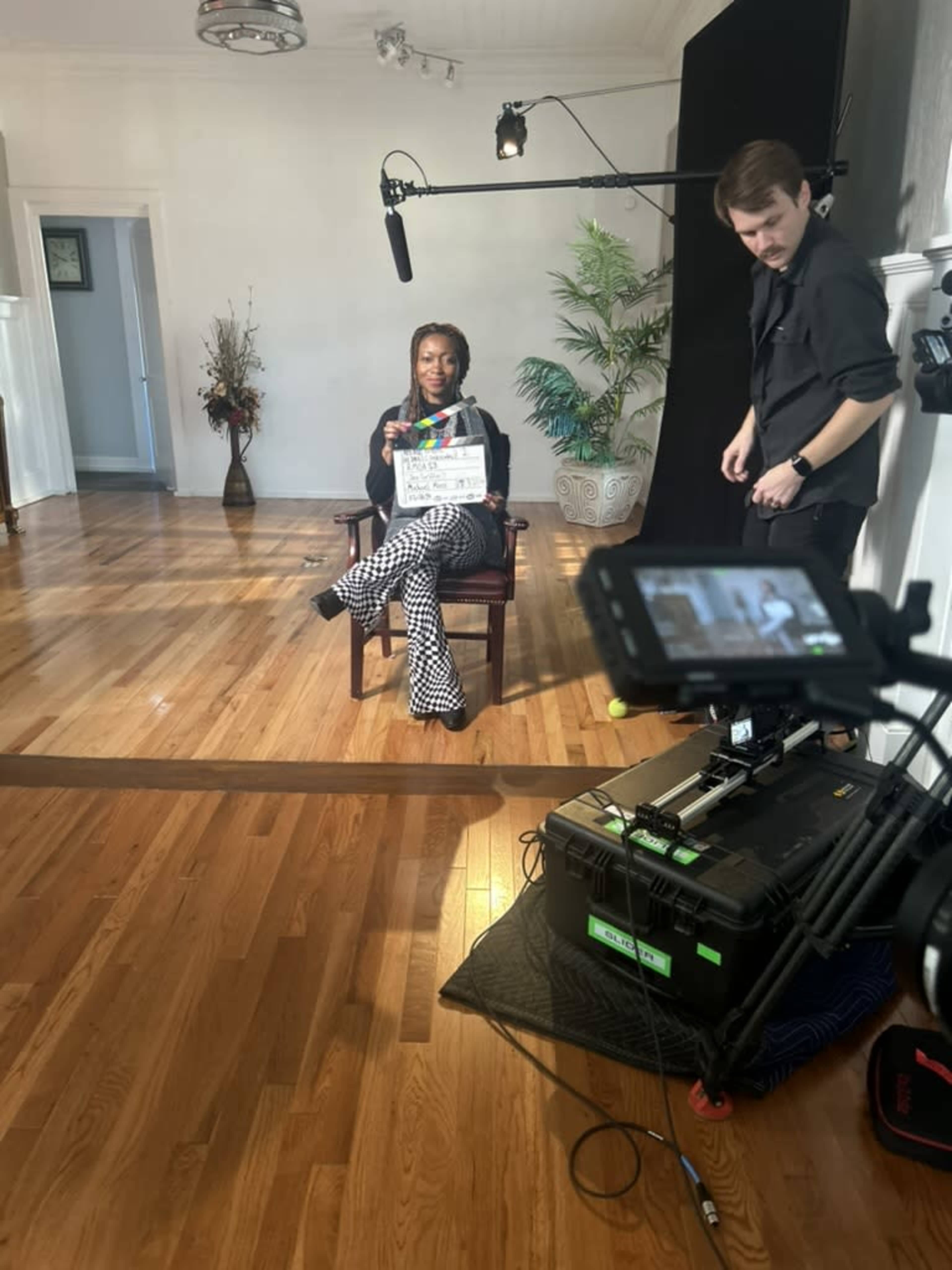 A woman sitting in a chair holds a clapperboard while a man operates a camera and microphone in a well-lit studio setting.