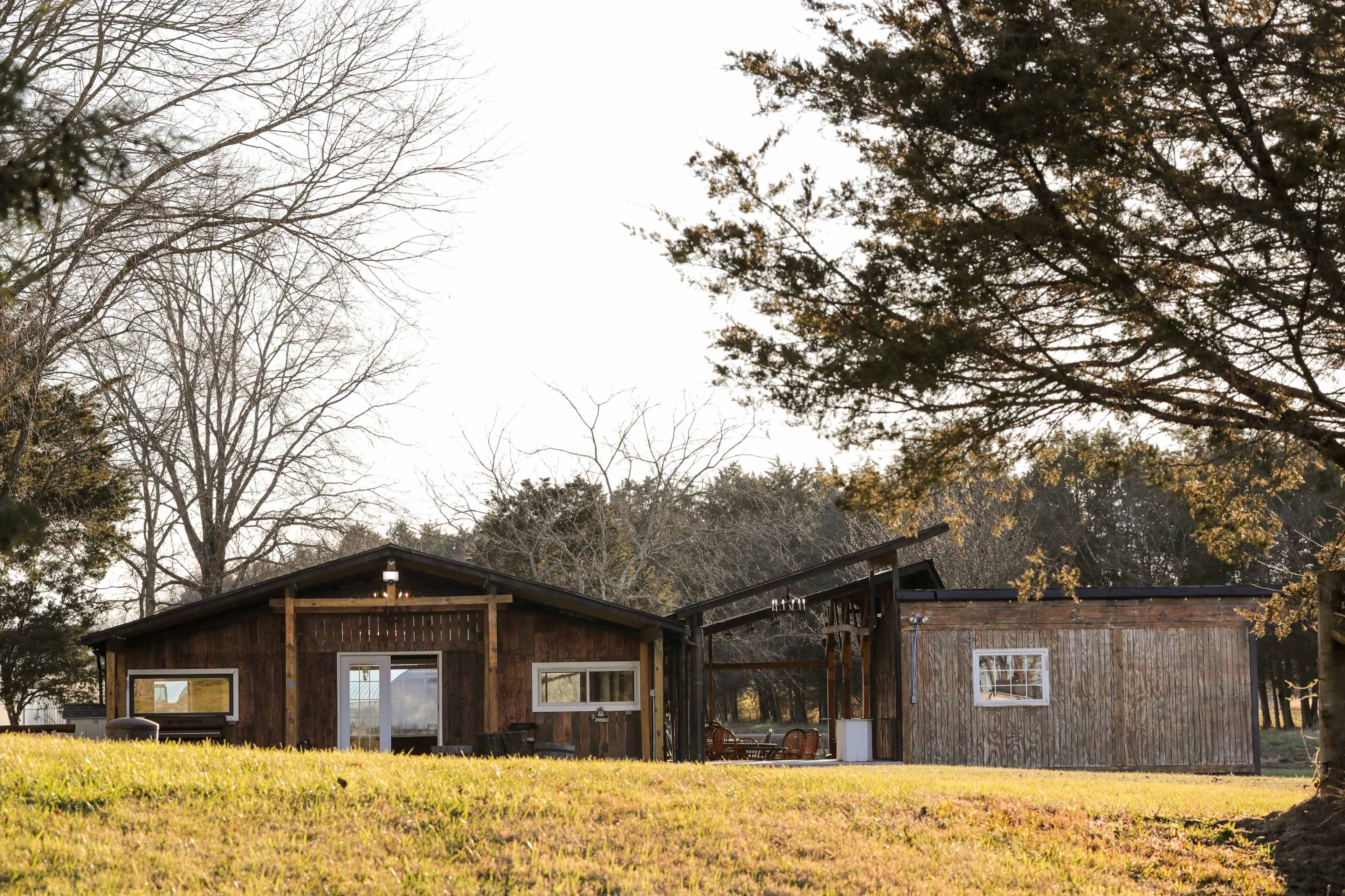 A rustic wooden house with a shed-style extension is situated on a grassy landscape surrounded by trees.