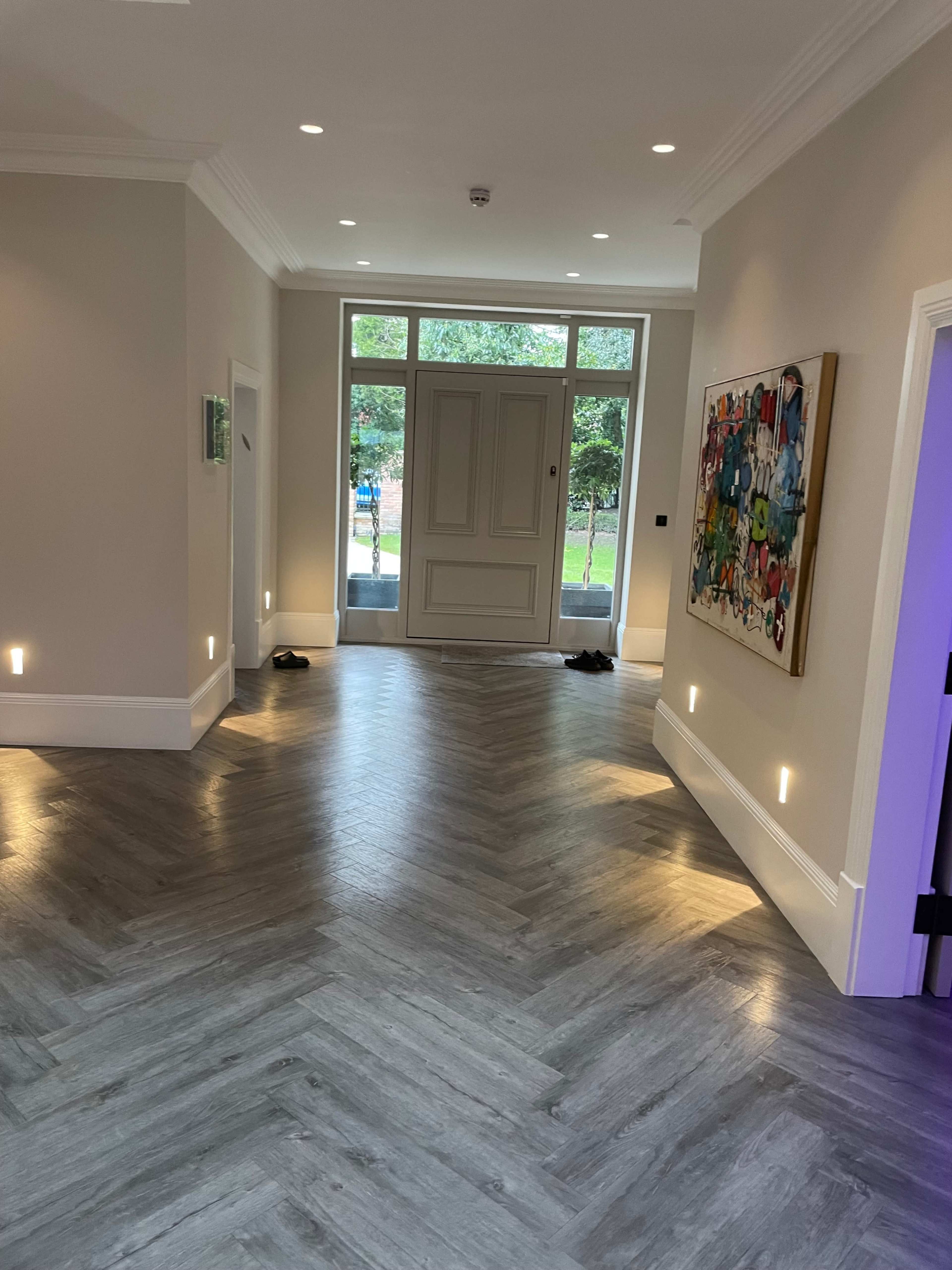The image shows a well-lit hallway with herringbone-patterned flooring, leading to a large entrance door framed by windows on either side.