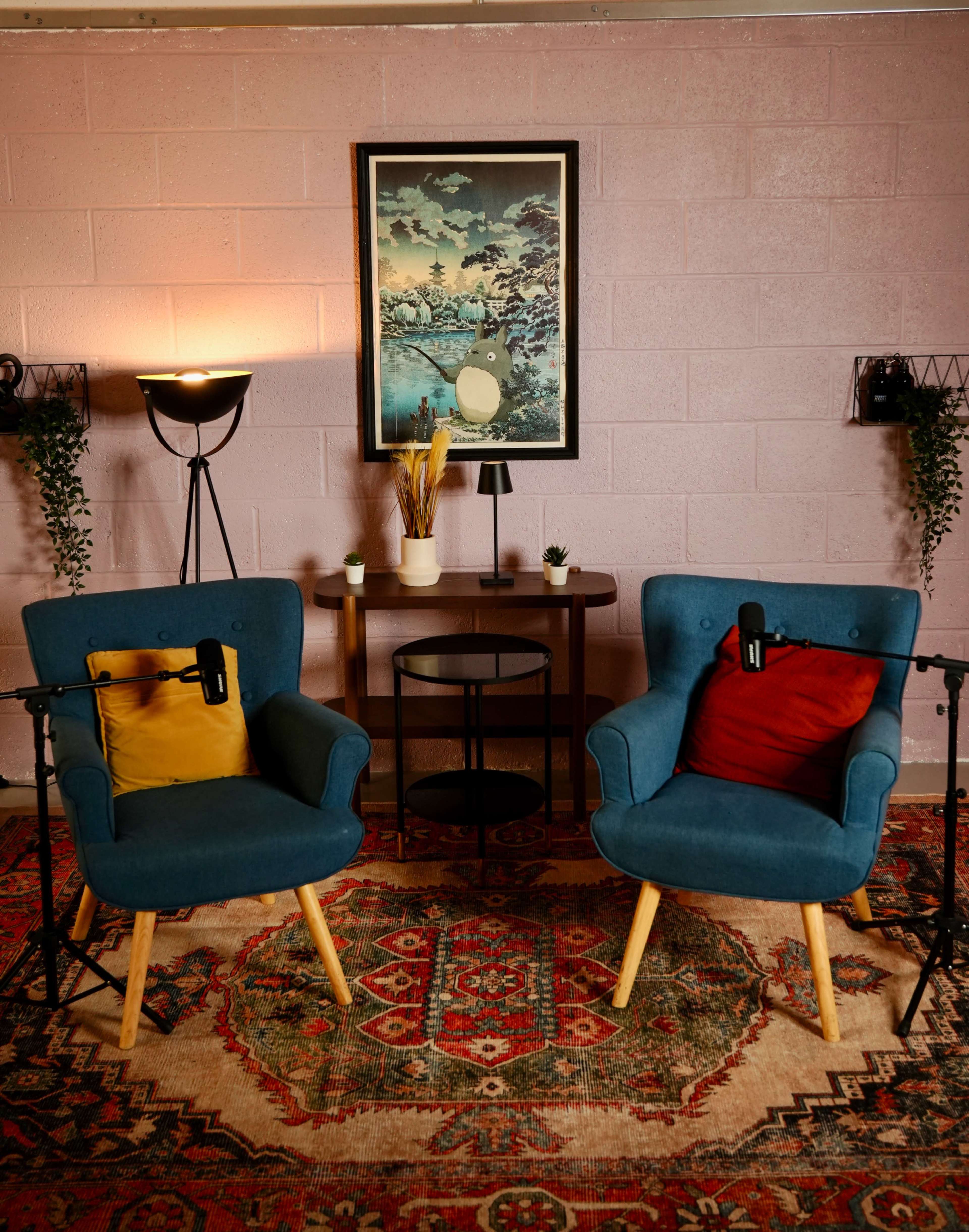 A cozy room features two blue armchairs with pillows, a circular table, and decorative plants against a pink wall.