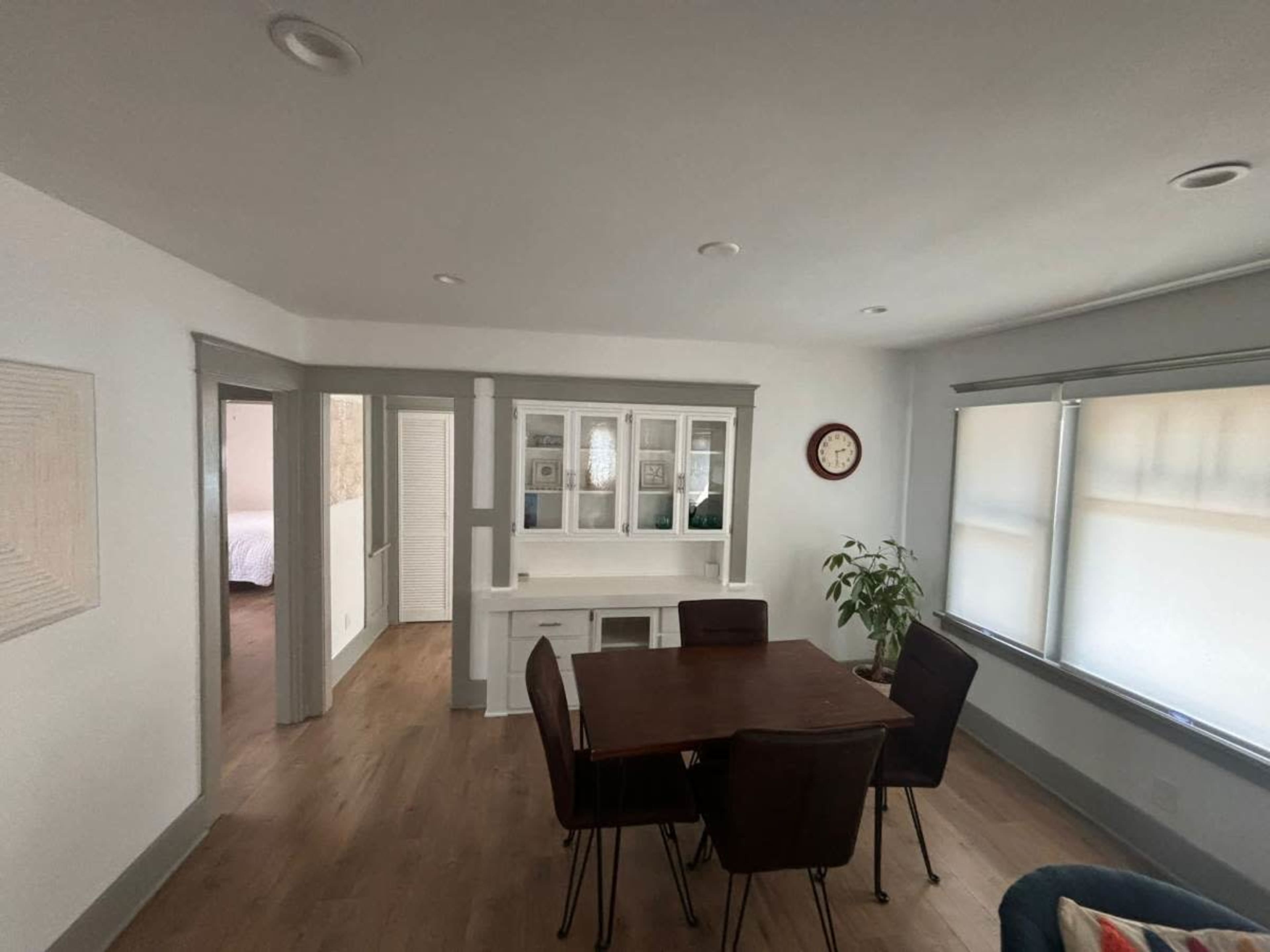 The image shows a dining area with a round table and six chairs, adjacent to a living space featuring a cabinet and large windows.