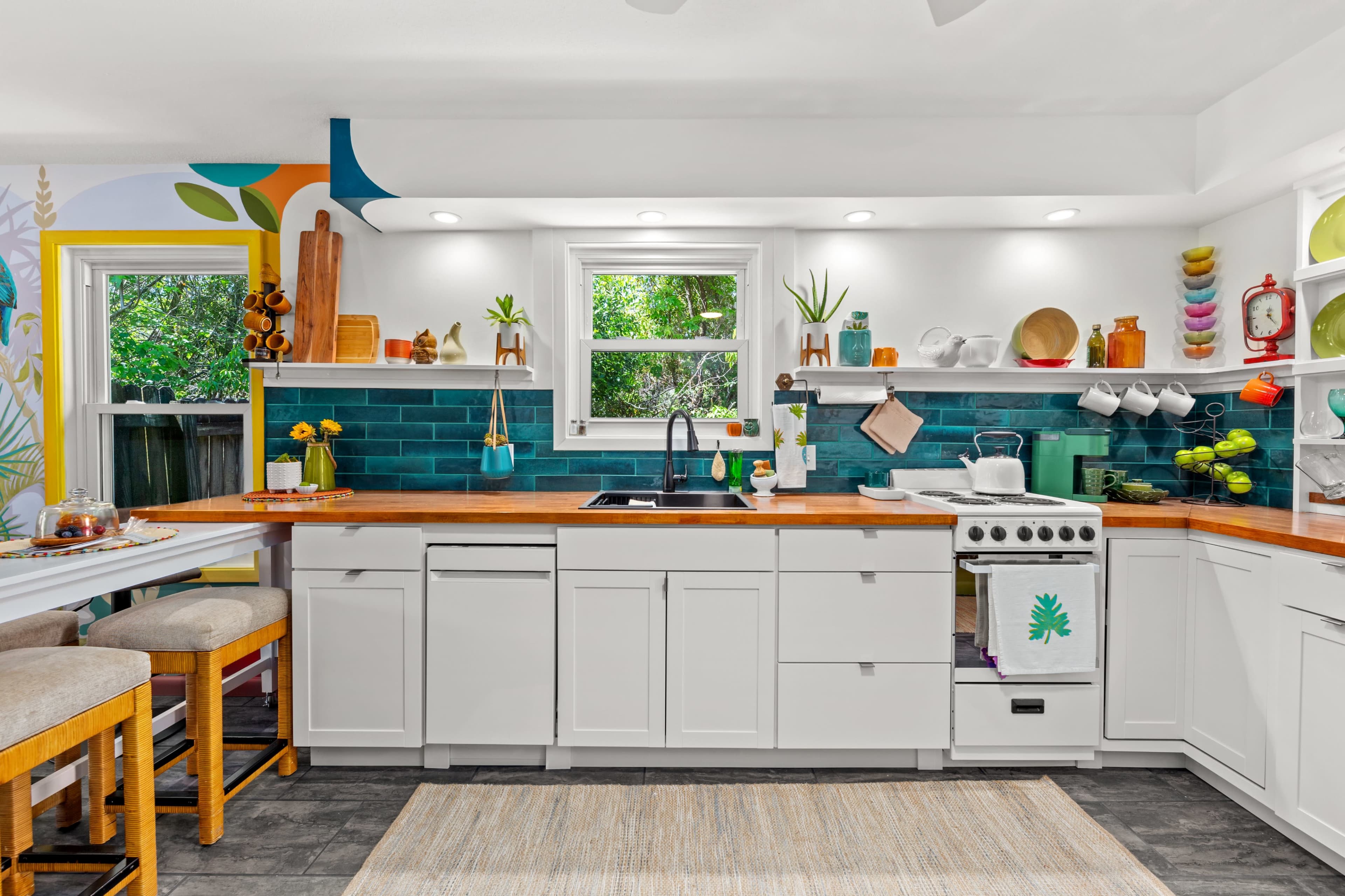 A modern kitchen with teal tiles, white cabinetry, a wooden countertop, and a variety of kitchenware arranged on open shelves.