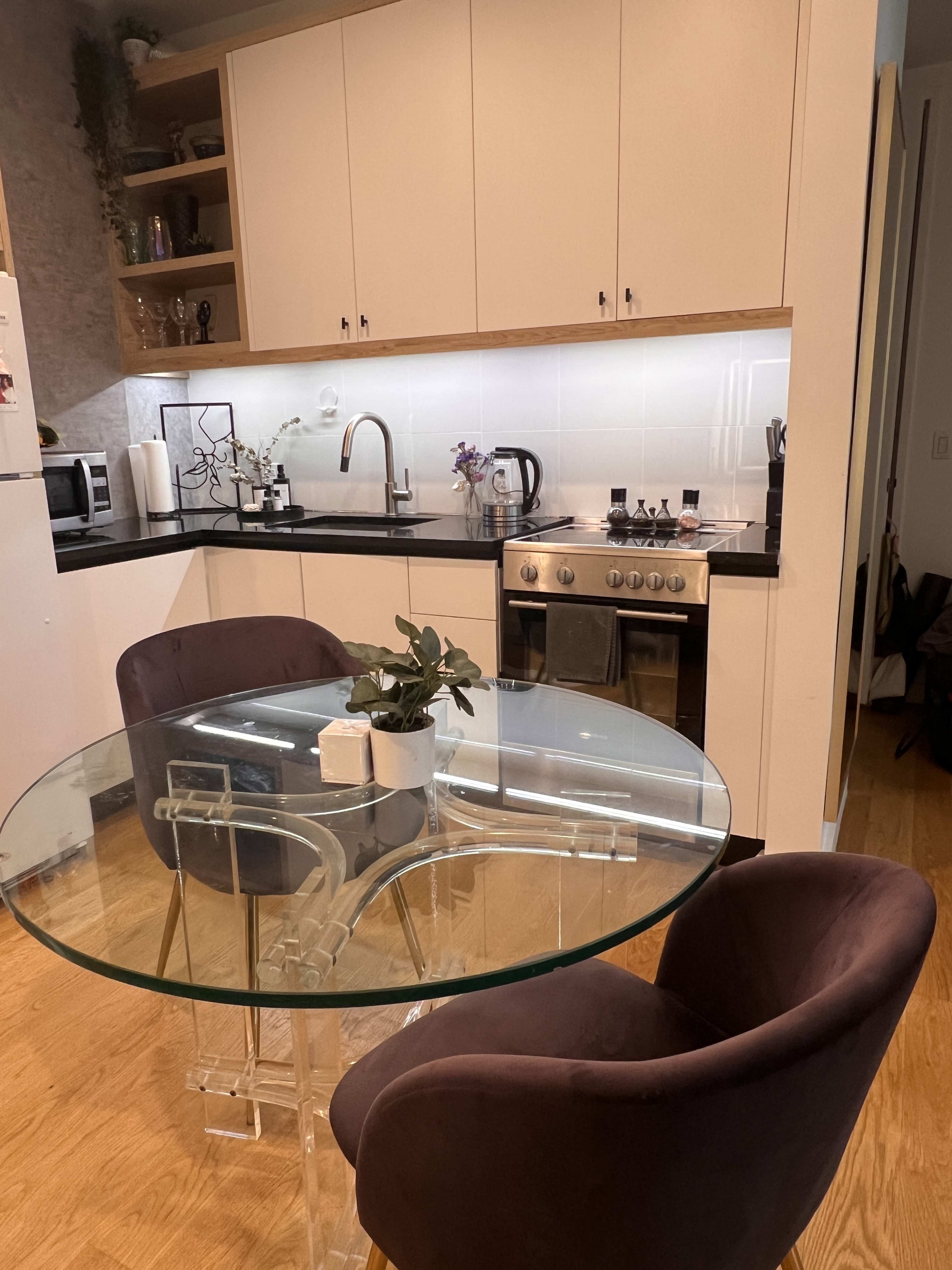 A modern kitchen with a glass dining table and two round velvet chairs, alongside stainless steel appliances and a white backsplash.