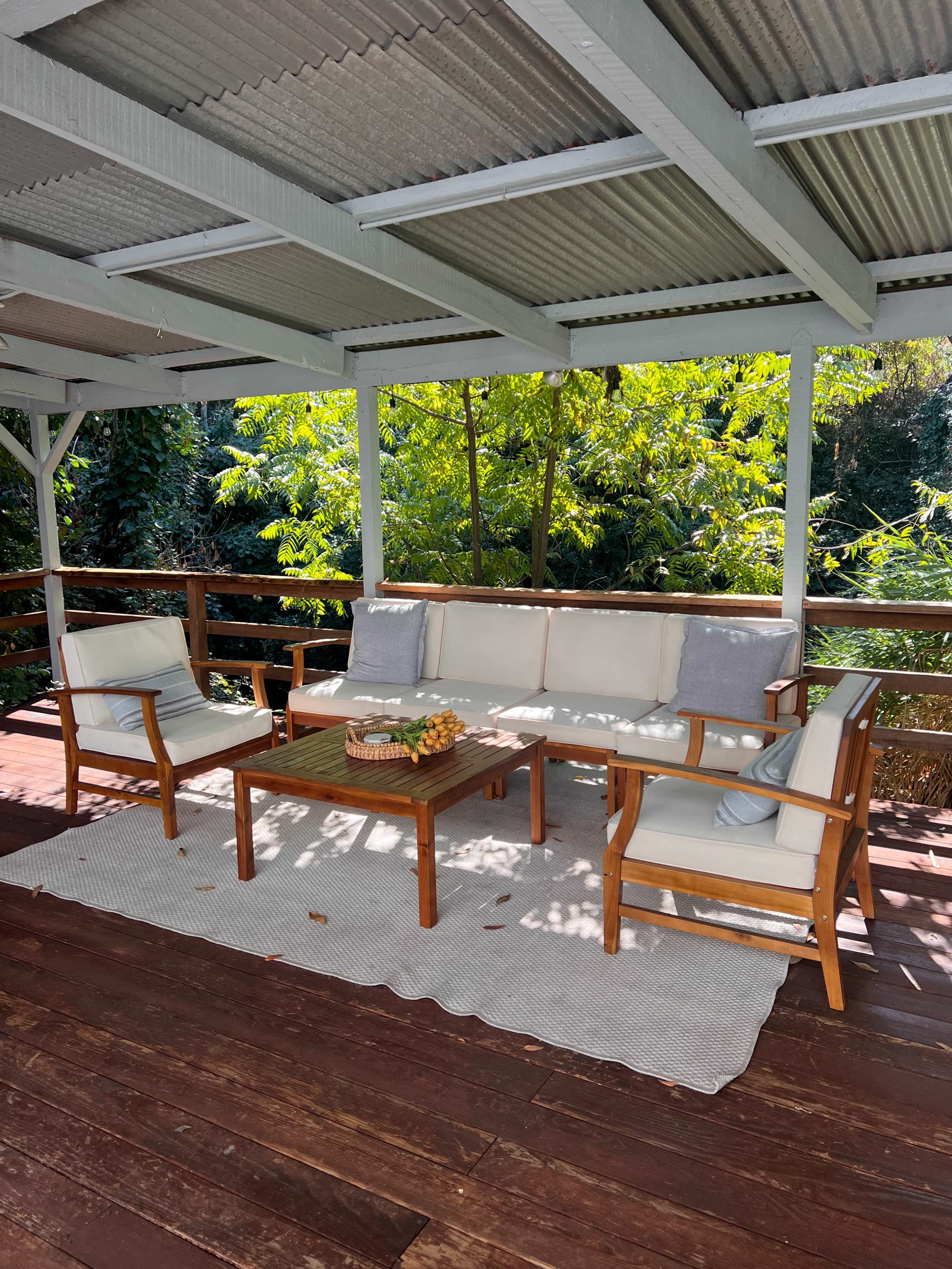 A covered outdoor seating area features a wooden table surrounded by four chairs and is adorned with a basket of fruit.