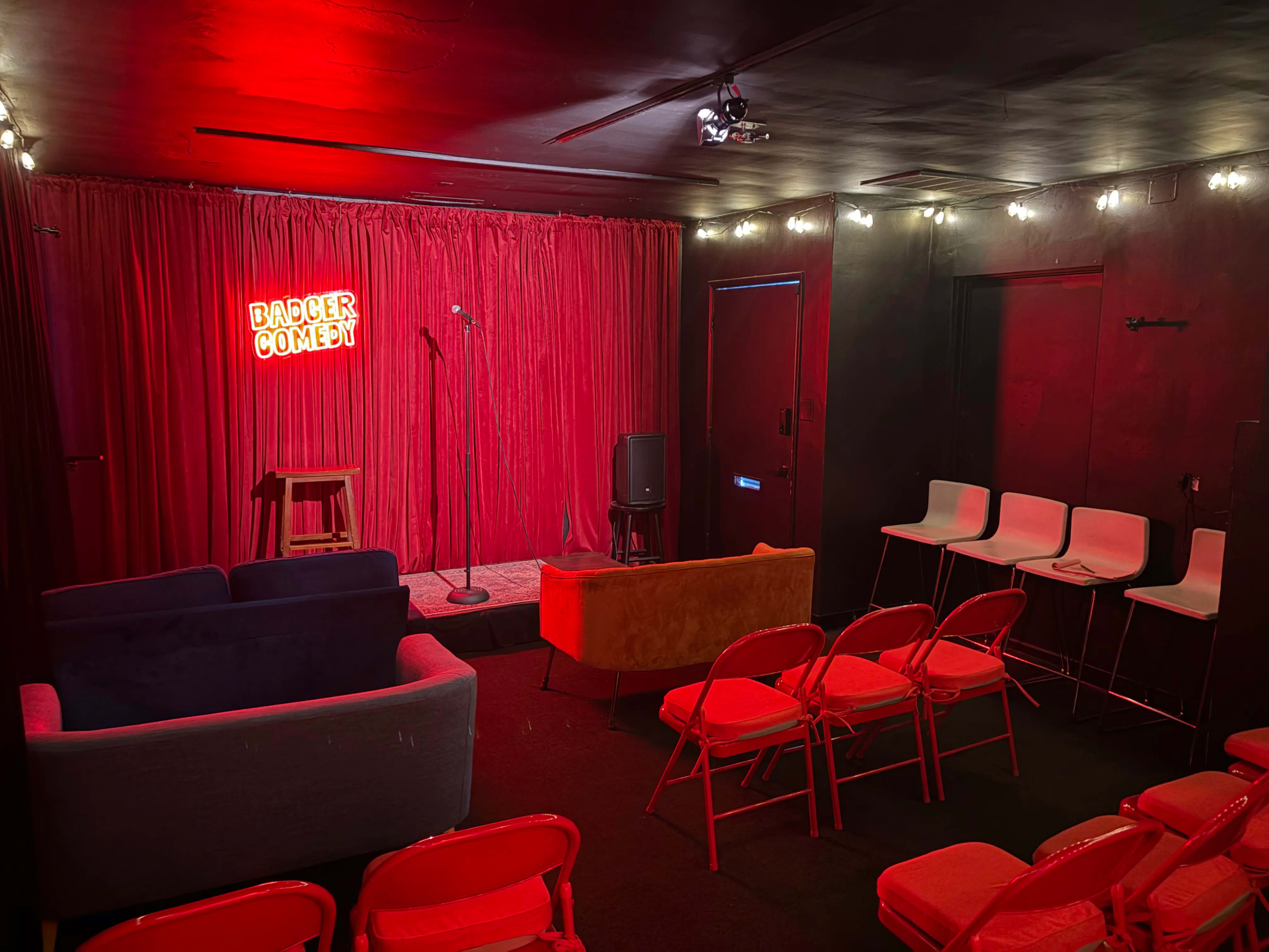 The image shows a small comedy venue with a red curtain backdrop, a microphone stand, and fabric seating arranged in rows.