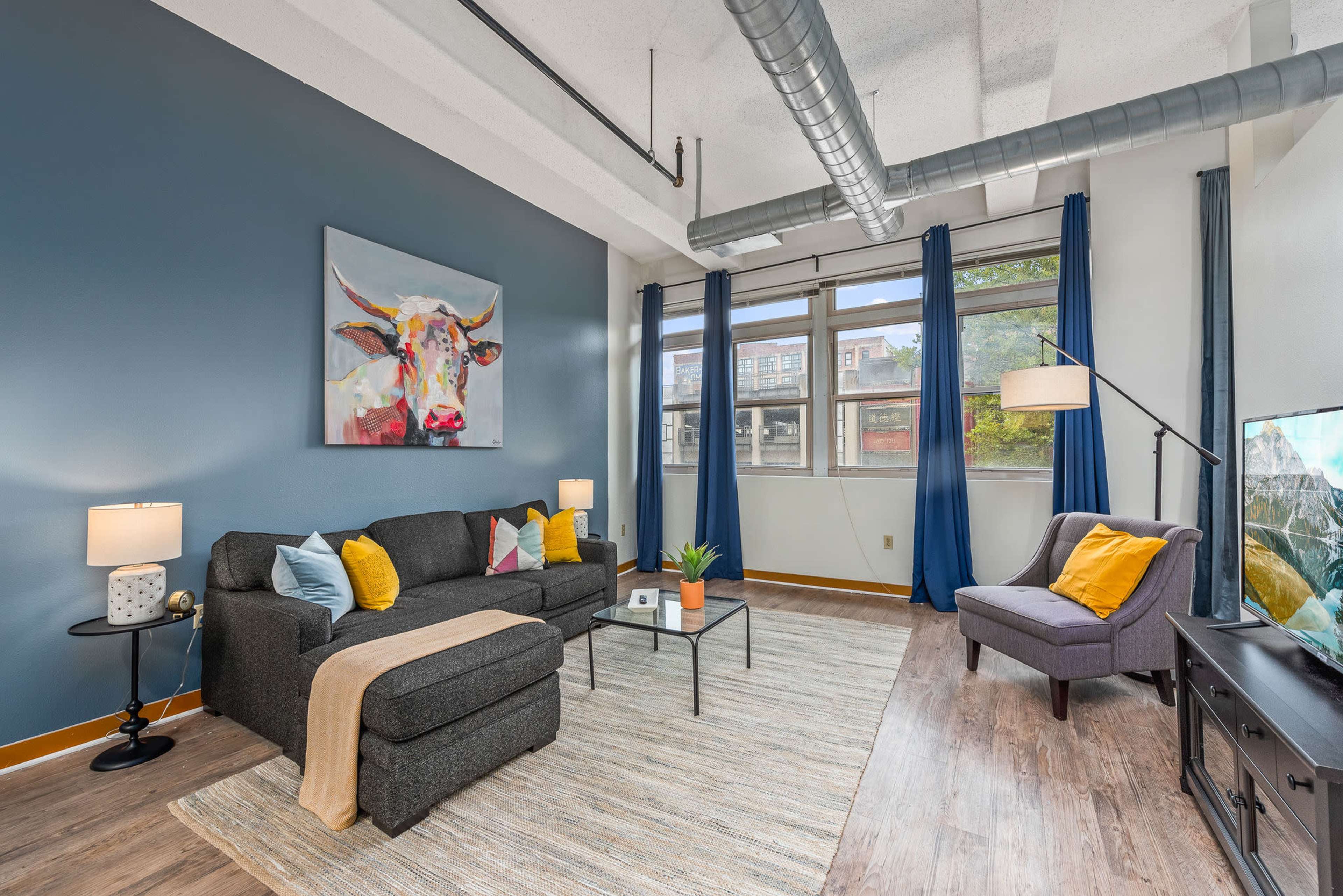 A modern living room with a gray sofa, yellow and blue accent pillows, a coffee table, and a large window with blue curtains, featuring a painting of a cow on the wall.