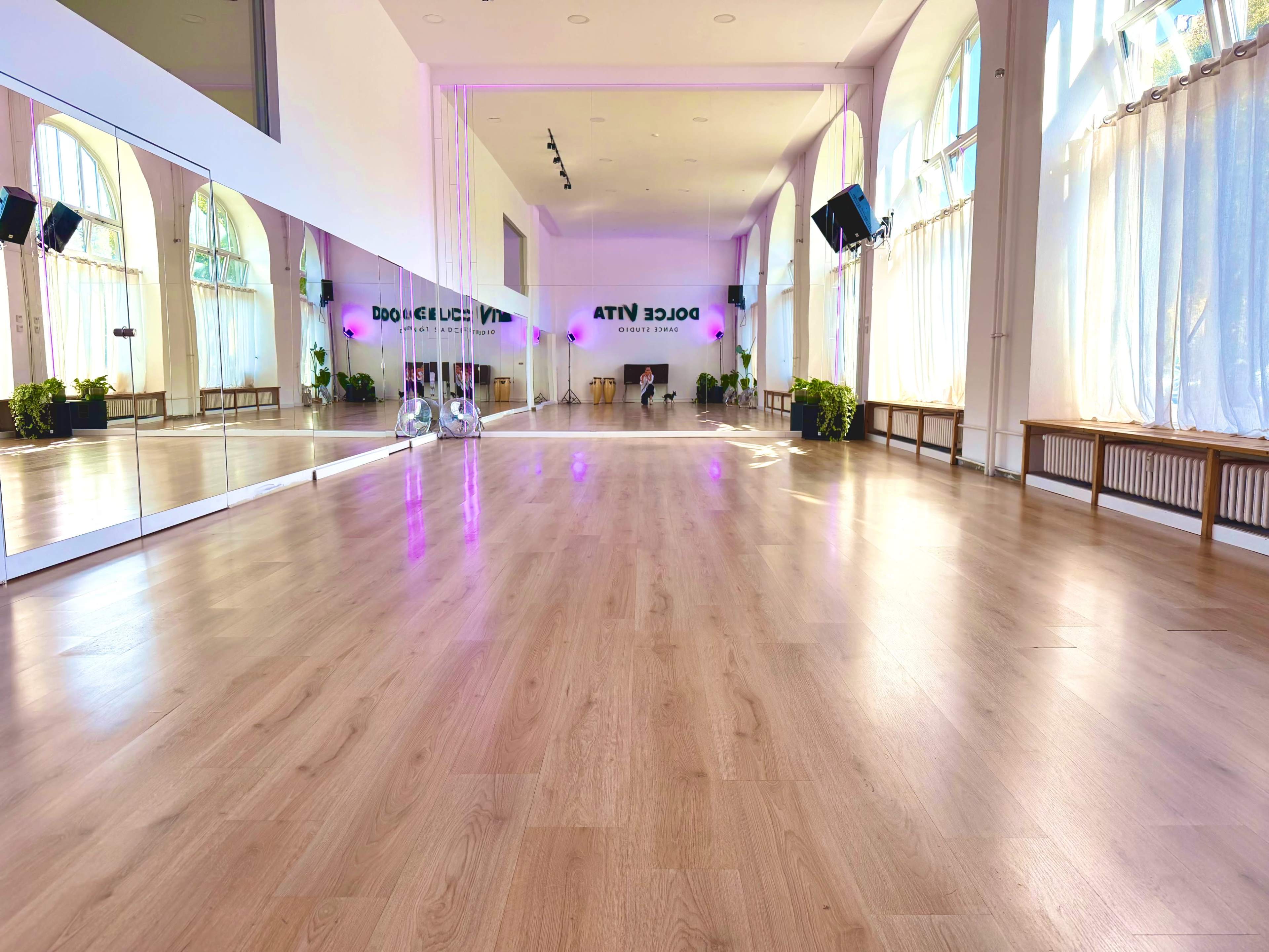 The image shows a bright dance studio with wooden floors, large windows, and mirrored walls, featuring plants and soft lighting.