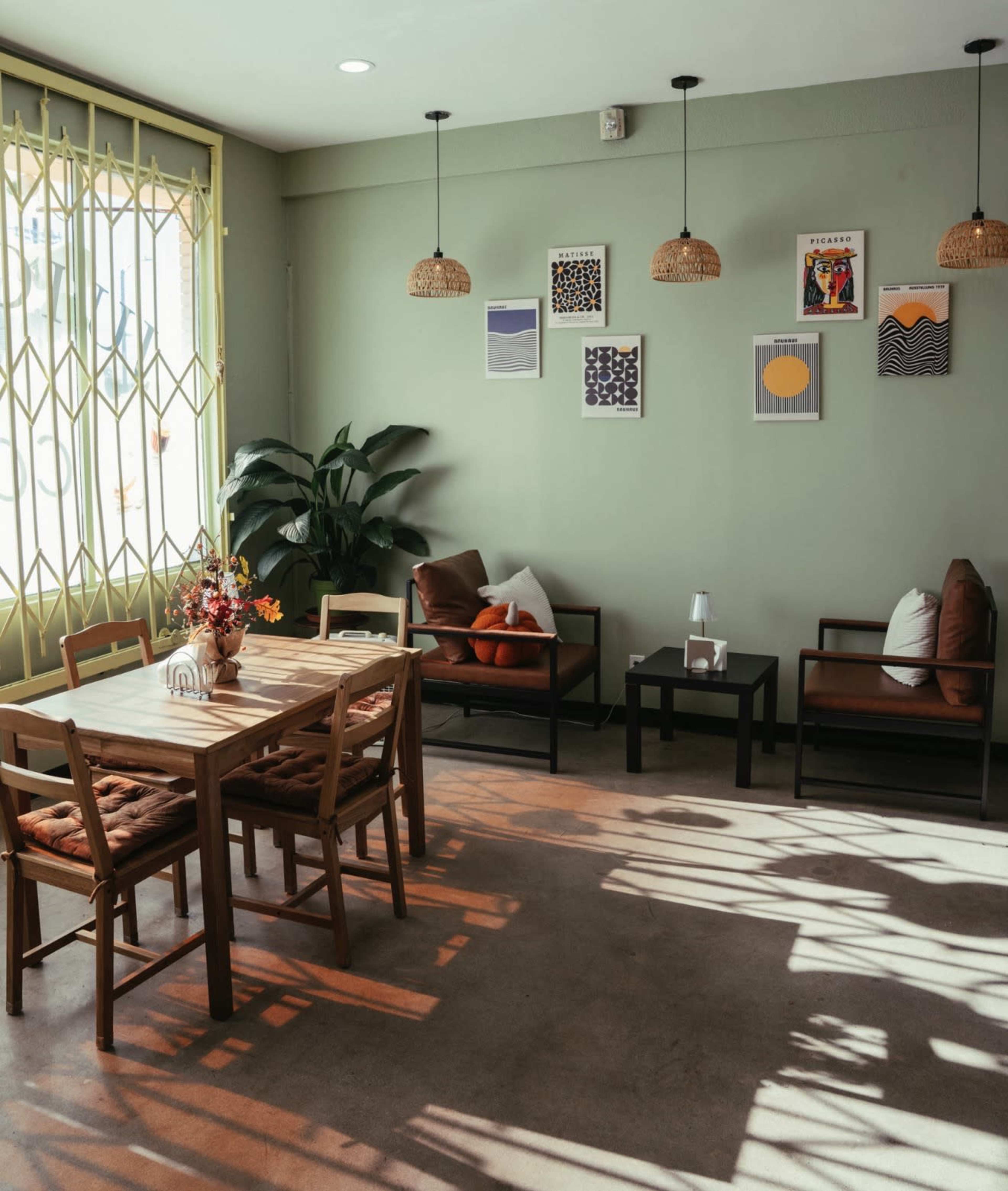 The image shows a cozy café interior with a wooden dining table and chairs, a few armchairs, green walls, and framed artwork hanging above.