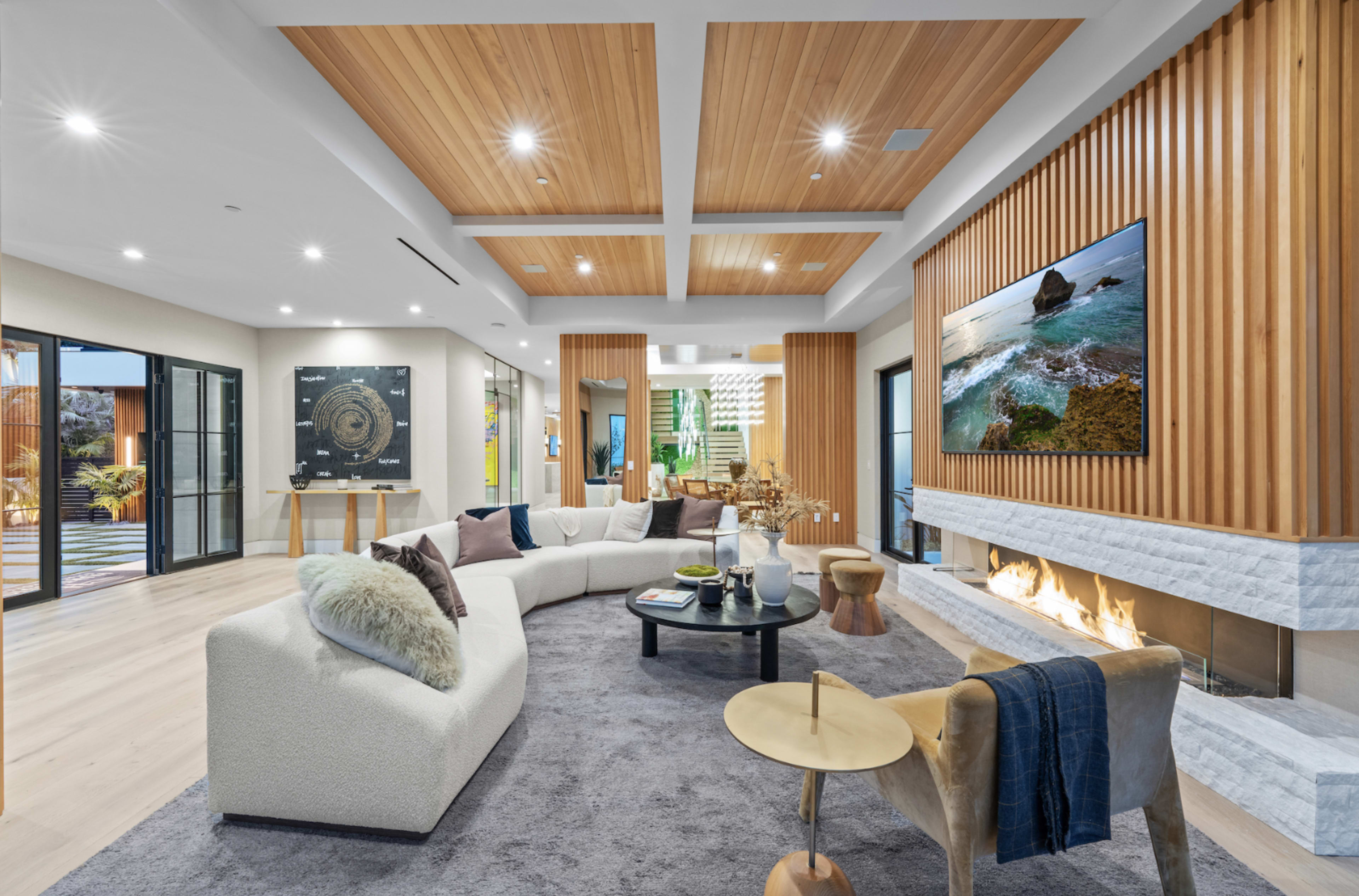 The image shows a modern living room with a curved sofa, a stone fireplace, and large windows featuring natural light and wooden ceiling beams.