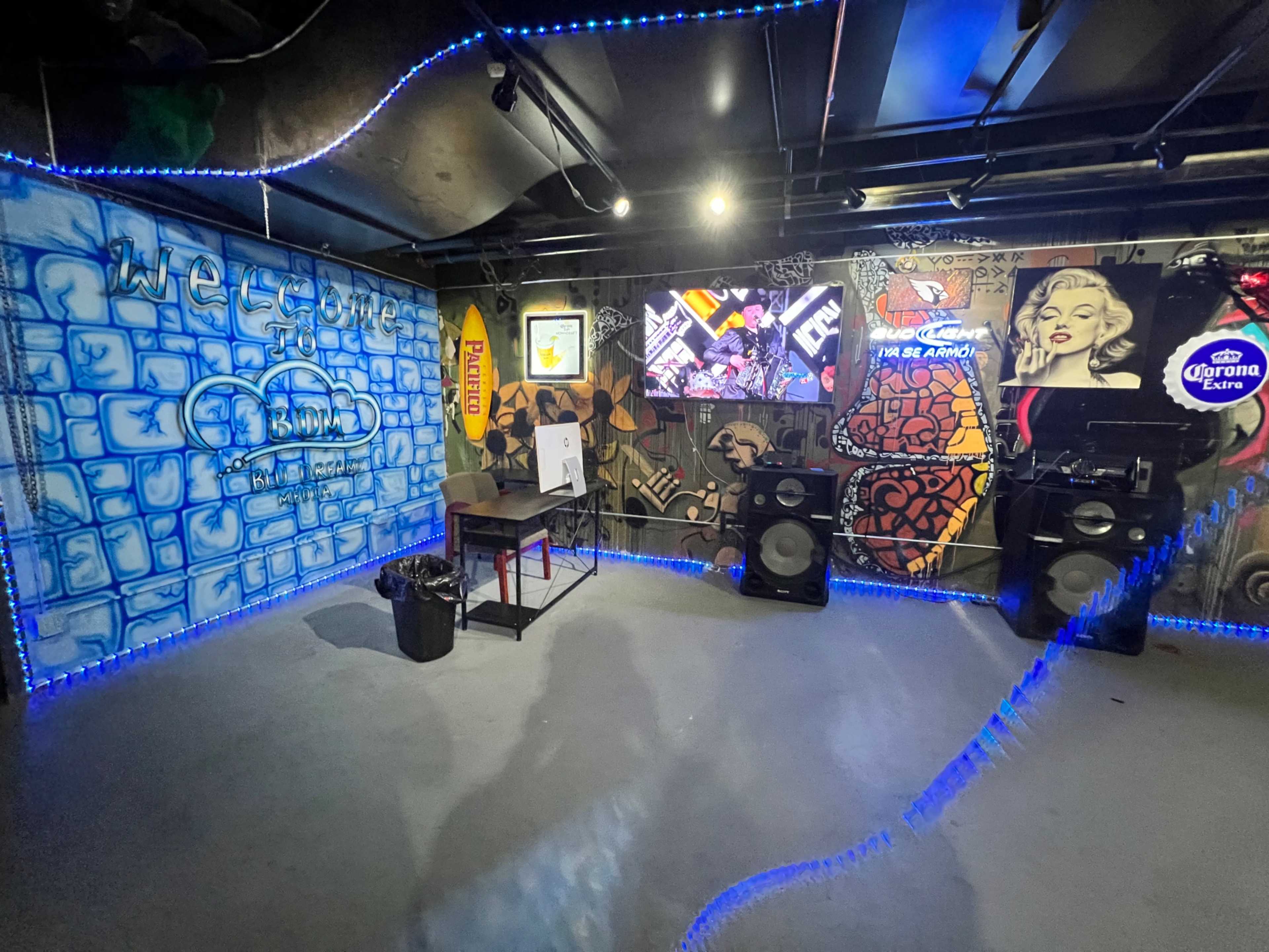 The image shows an underground room with colorful graffiti on the walls, a table and a trash can on the floor, and large speakers positioned near a television screen.