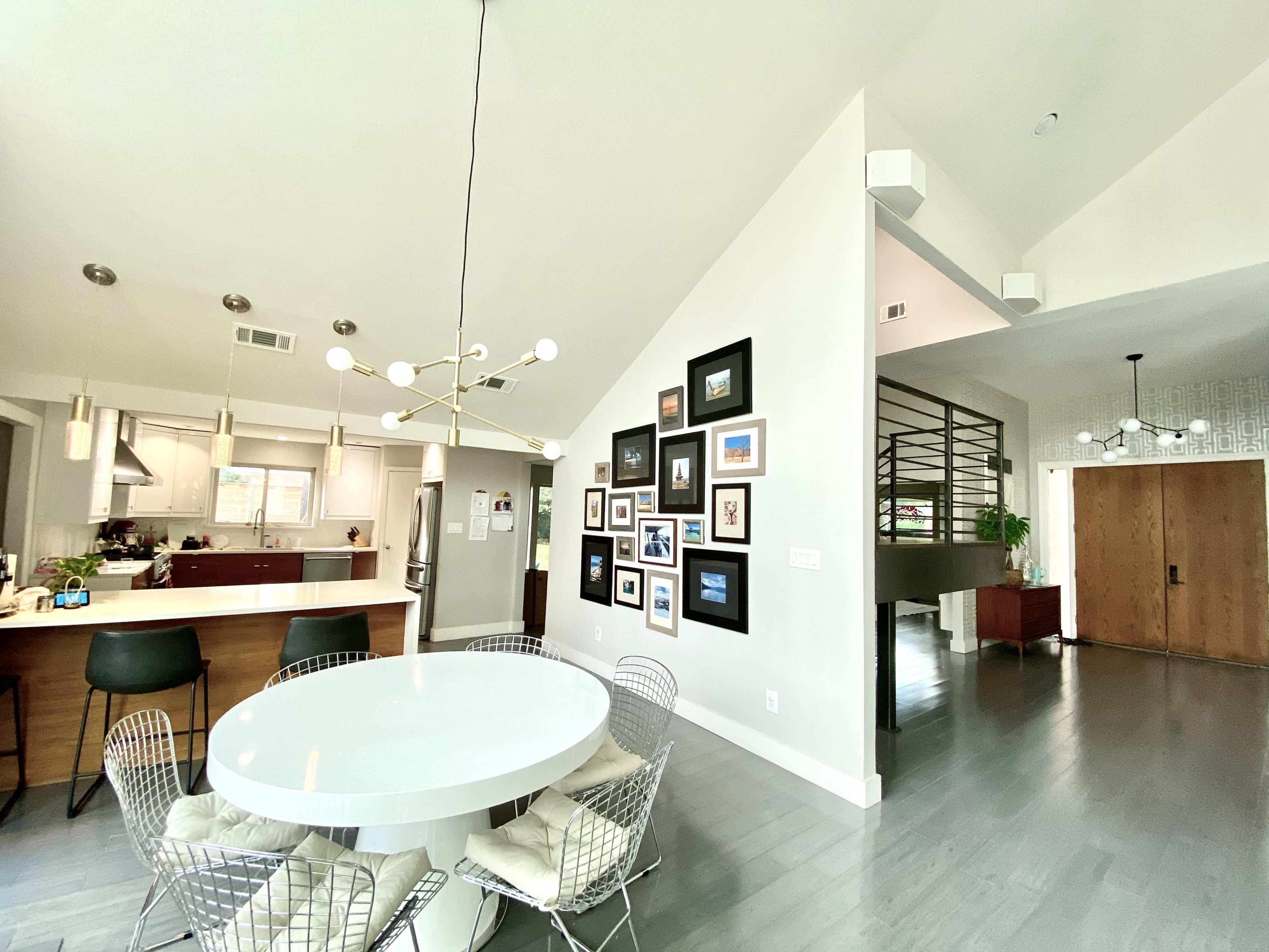 A spacious, modern home interior features a round white dining table with wire chairs, a gallery wall of framed pictures, and a large open kitchen visible in the background.