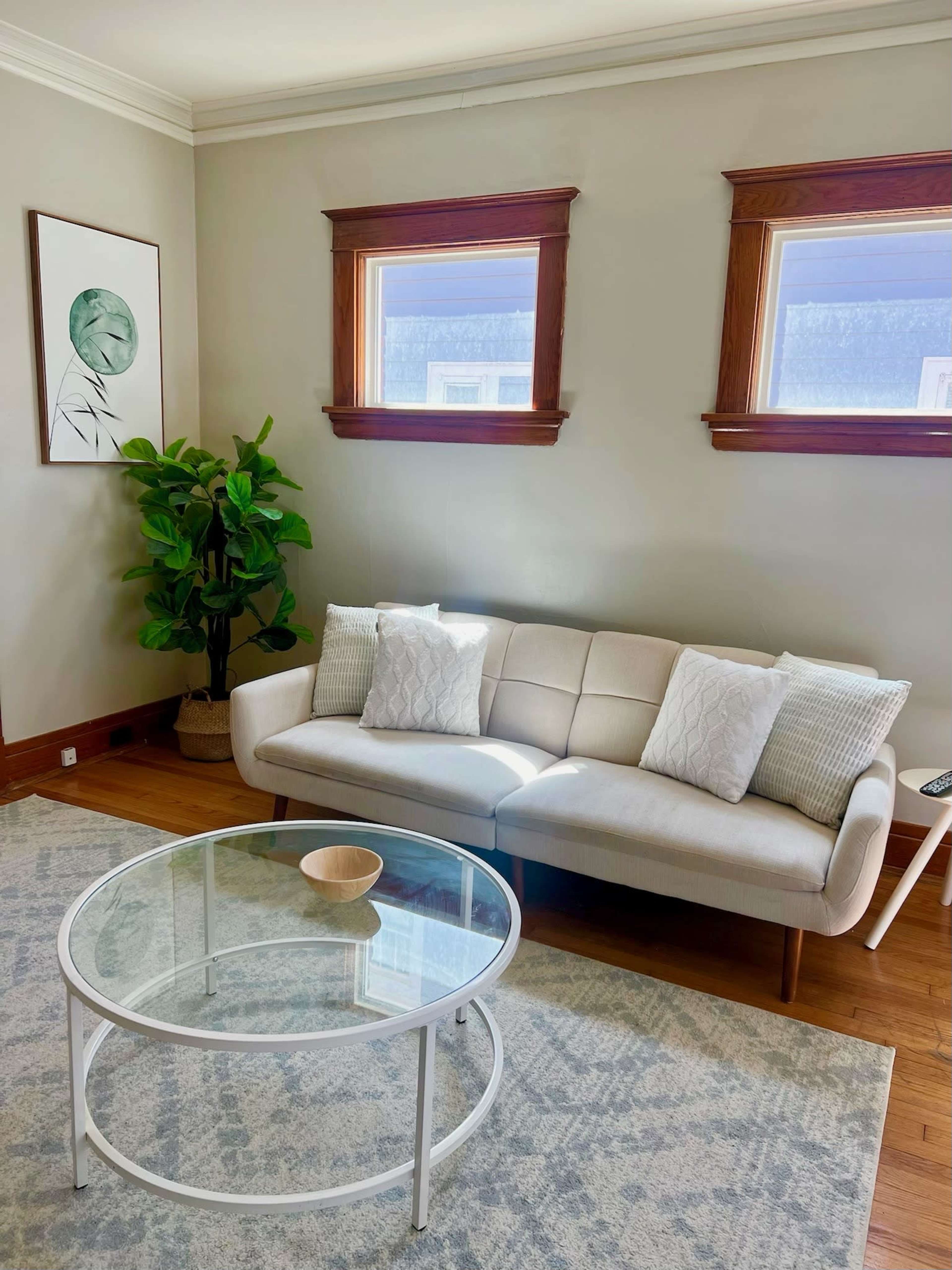 A light-filled living room features a white sofa with decorative pillows, a round glass coffee table, and a potted plant beside two windows with wooden frames.