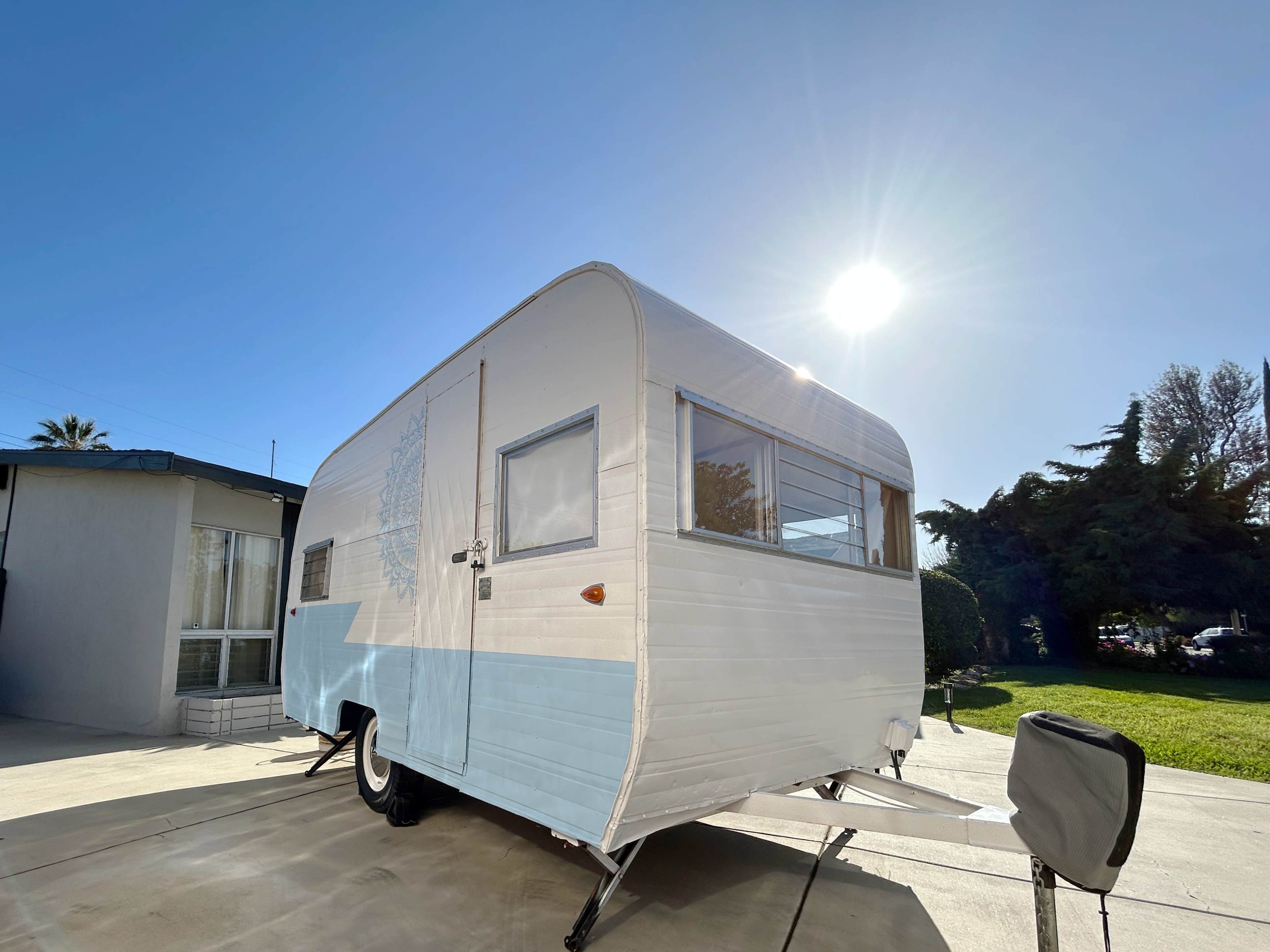 A vintage camper trailer is parked on a concrete driveway with the sun shining brightly overhead.