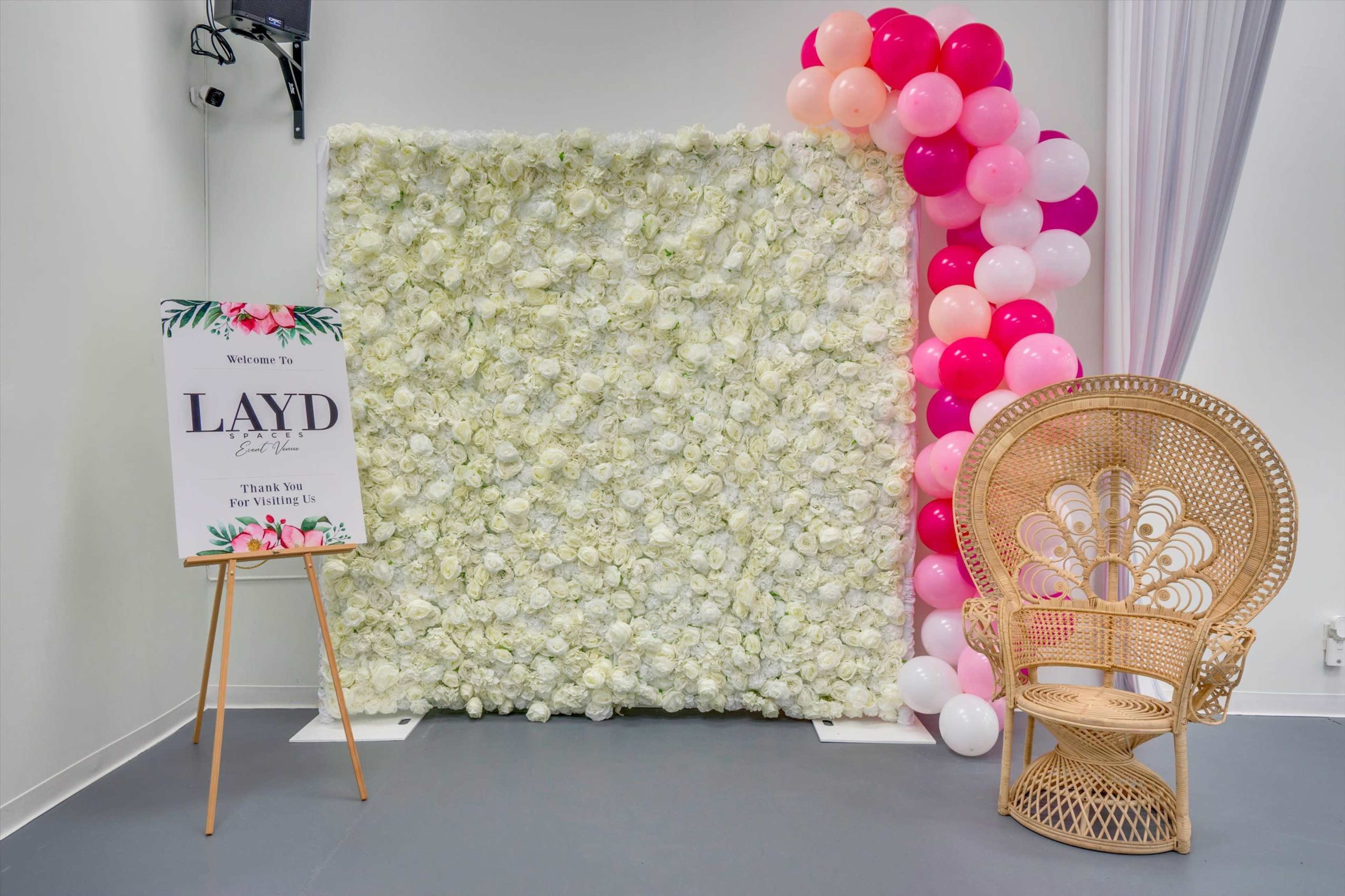 The image features a white floral backdrop with a colorful balloon arrangement on one side and a decorative wicker chair in front of it.