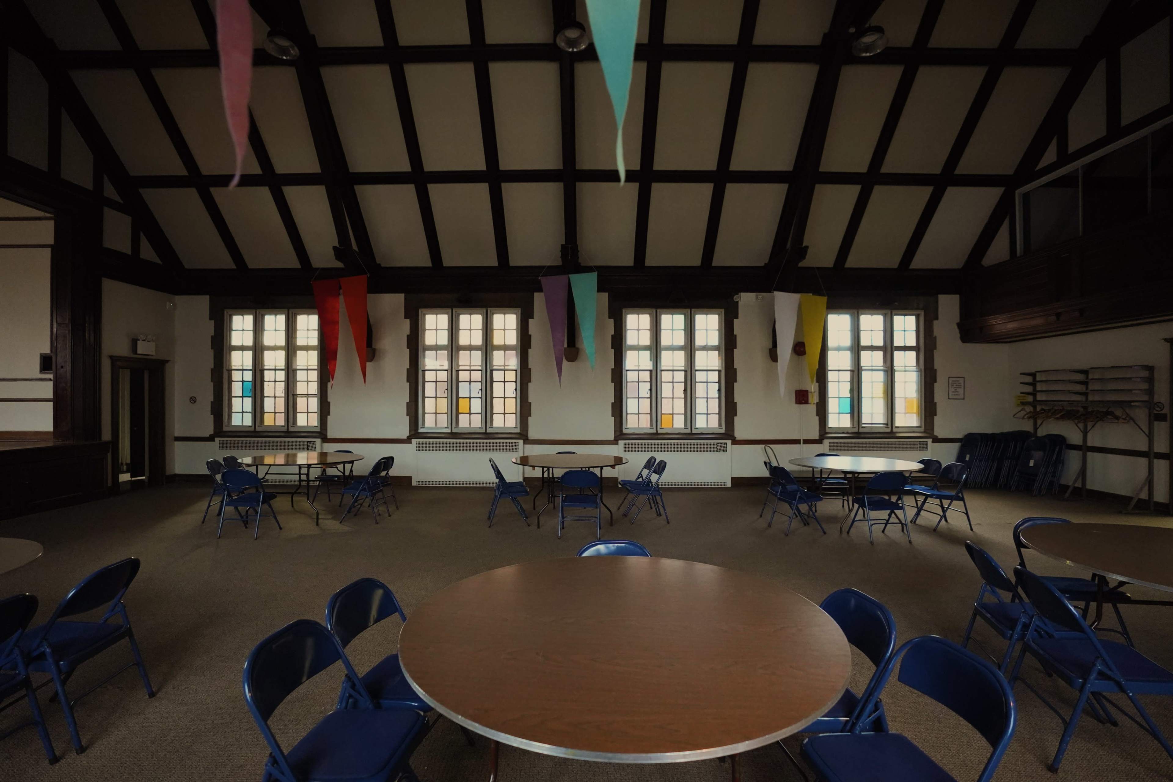 A large, empty room features several round tables surrounded by blue chairs, with colorful banners hanging from the wooden ceiling and windows lining the walls.