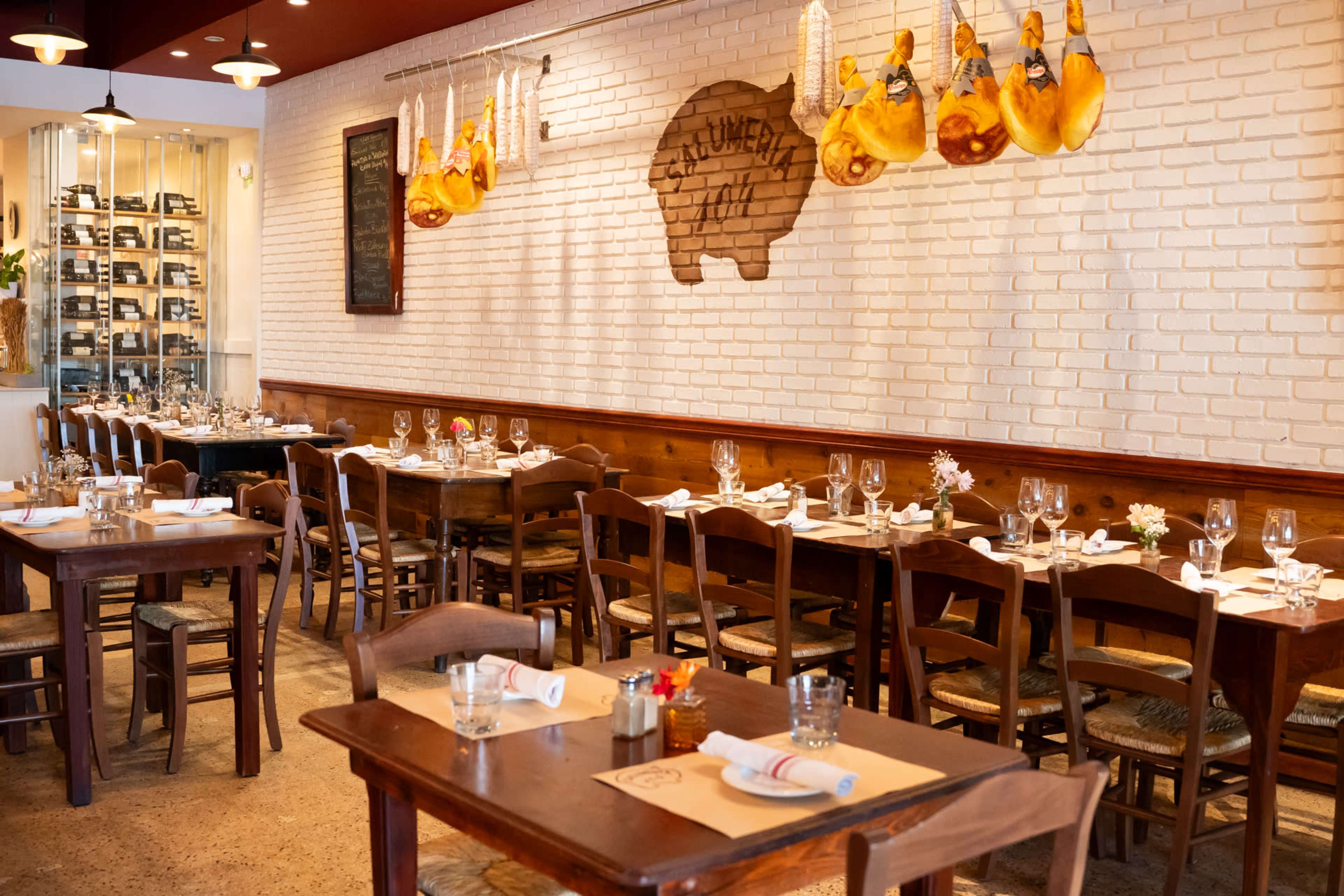 The image shows a restaurant interior with wooden tables and chairs arranged neatly, white brick walls, and hanging cured hams decoratively displayed.