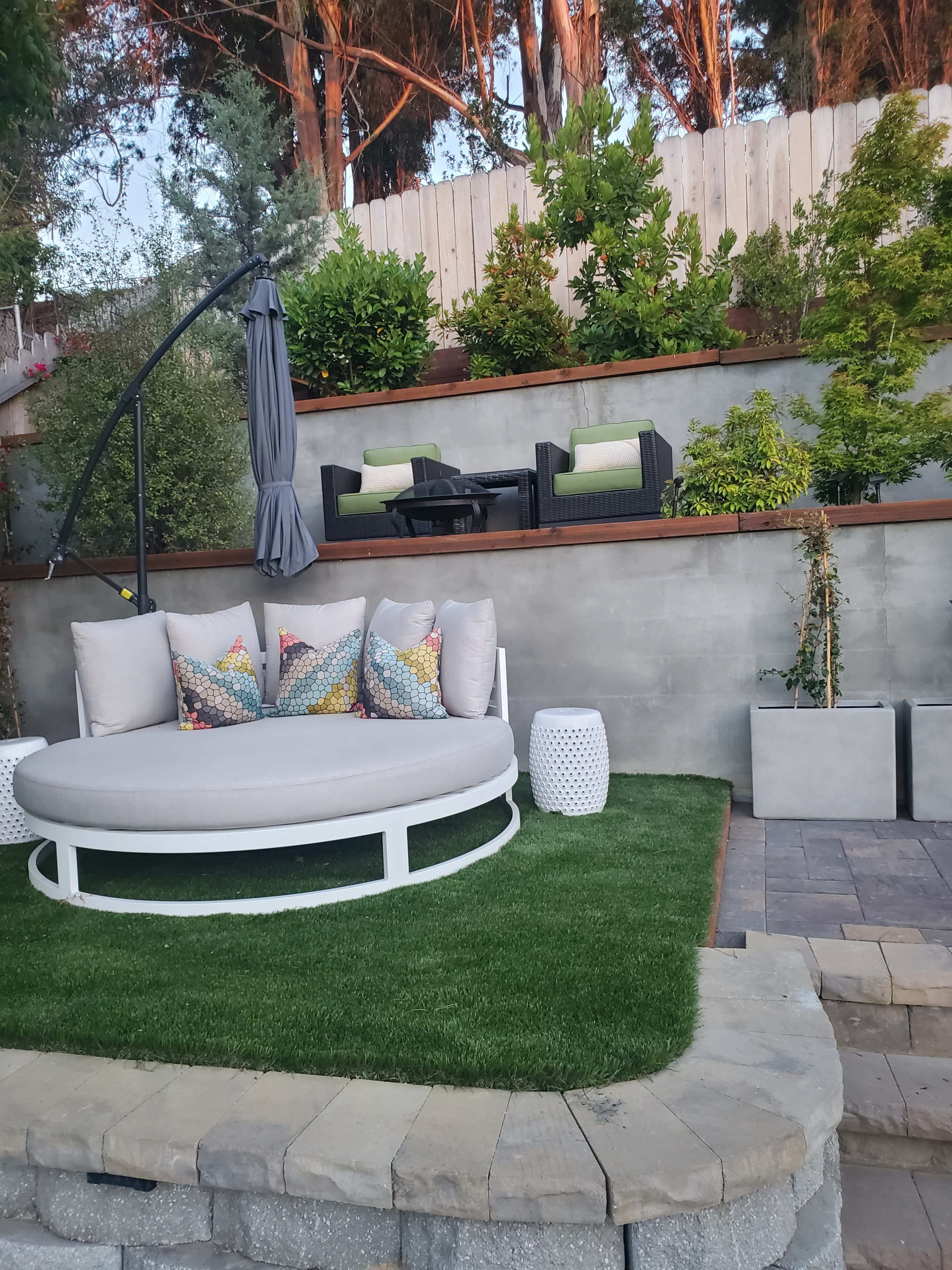 A landscaped backyard featuring a circular gray lounge bed with decorative pillows, surrounded by a small green lawn, stone paving, and seating arrangements on an elevated wooden deck.