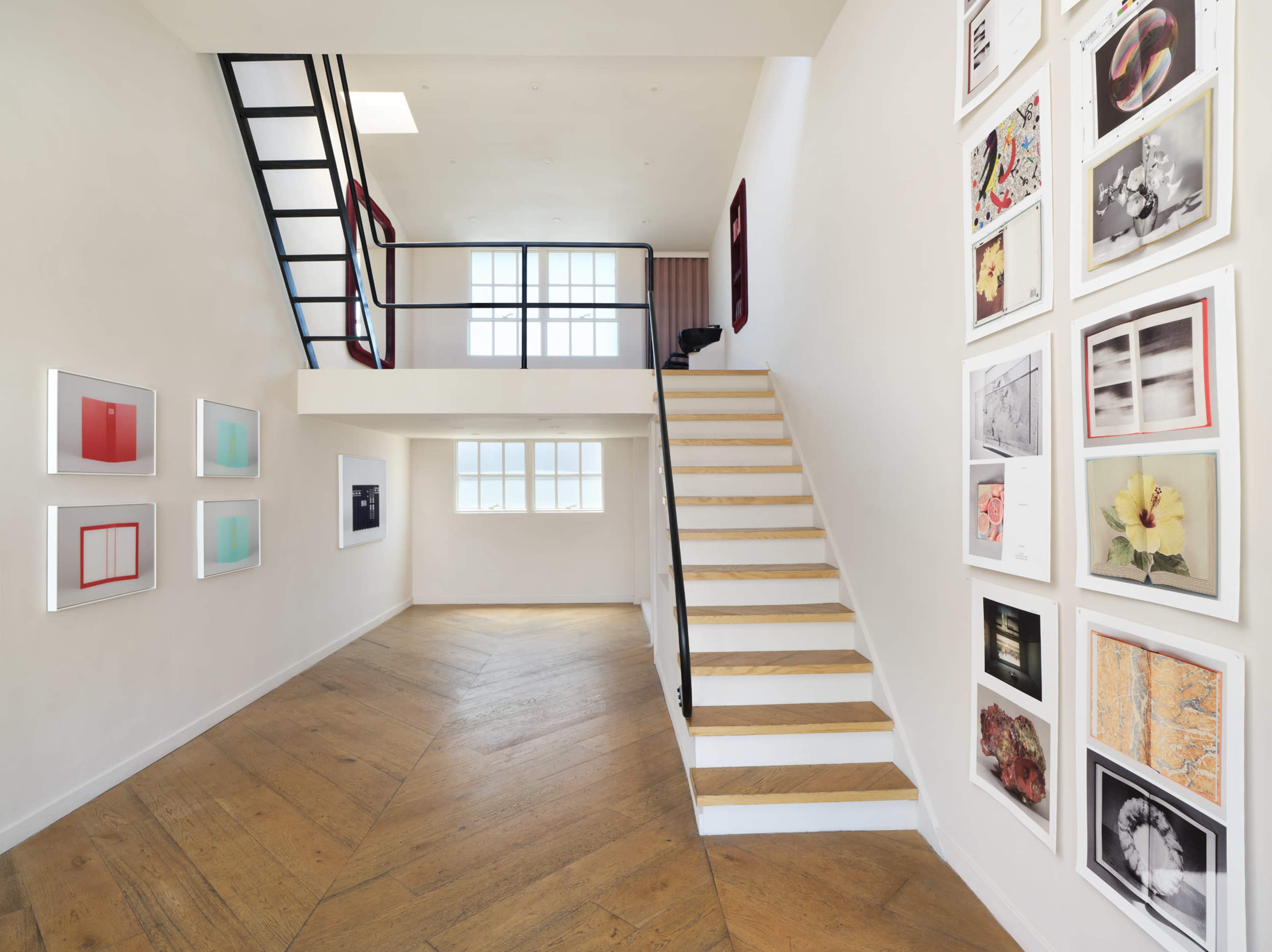 The image shows a modern interior with a staircase on the right, framed artwork on the left wall, and a large window allowing natural light to enter.