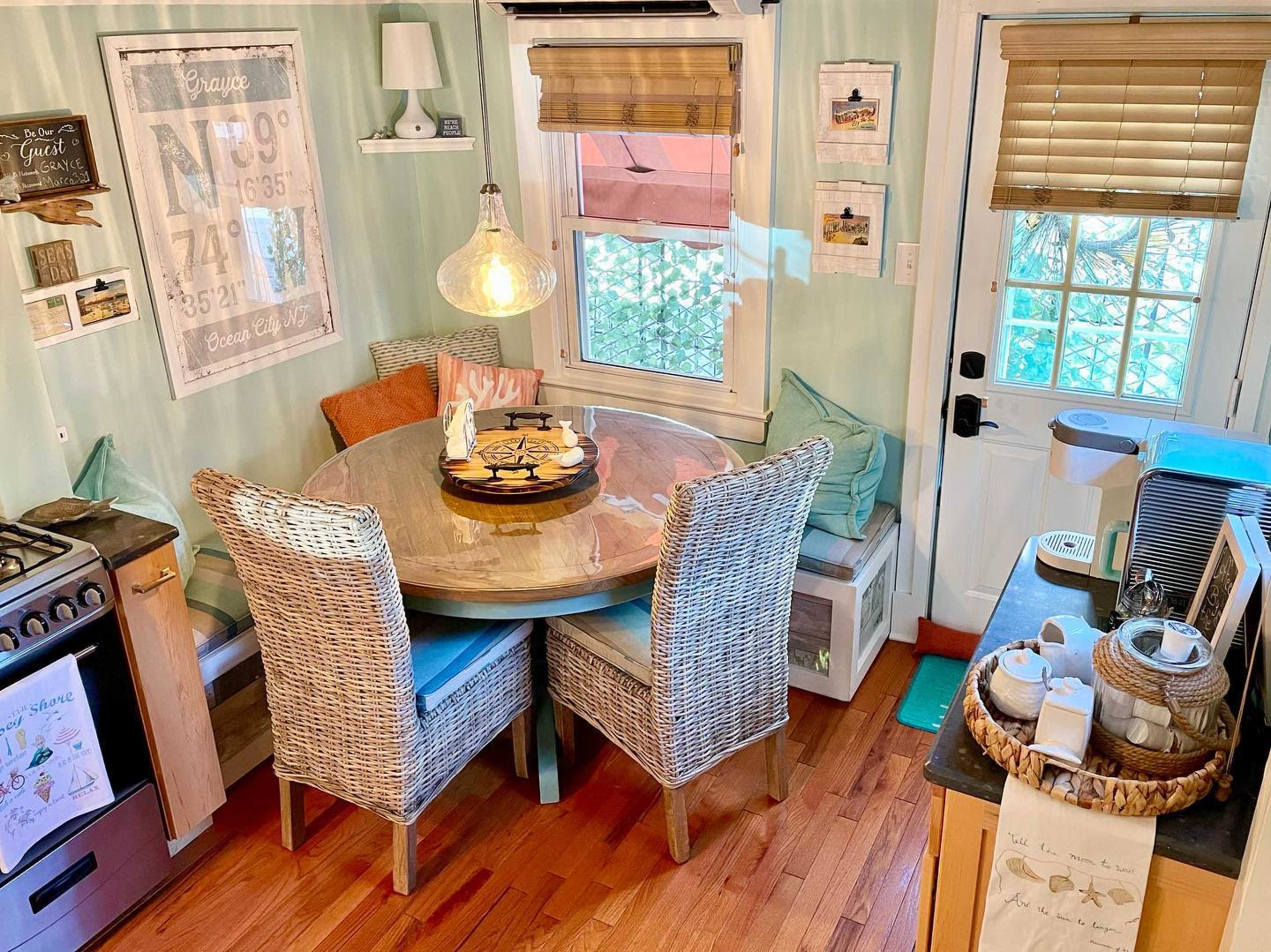 A cozy kitchen nook features a round table surrounded by four wicker chairs, with natural light filtering through the window and decor lining the walls.