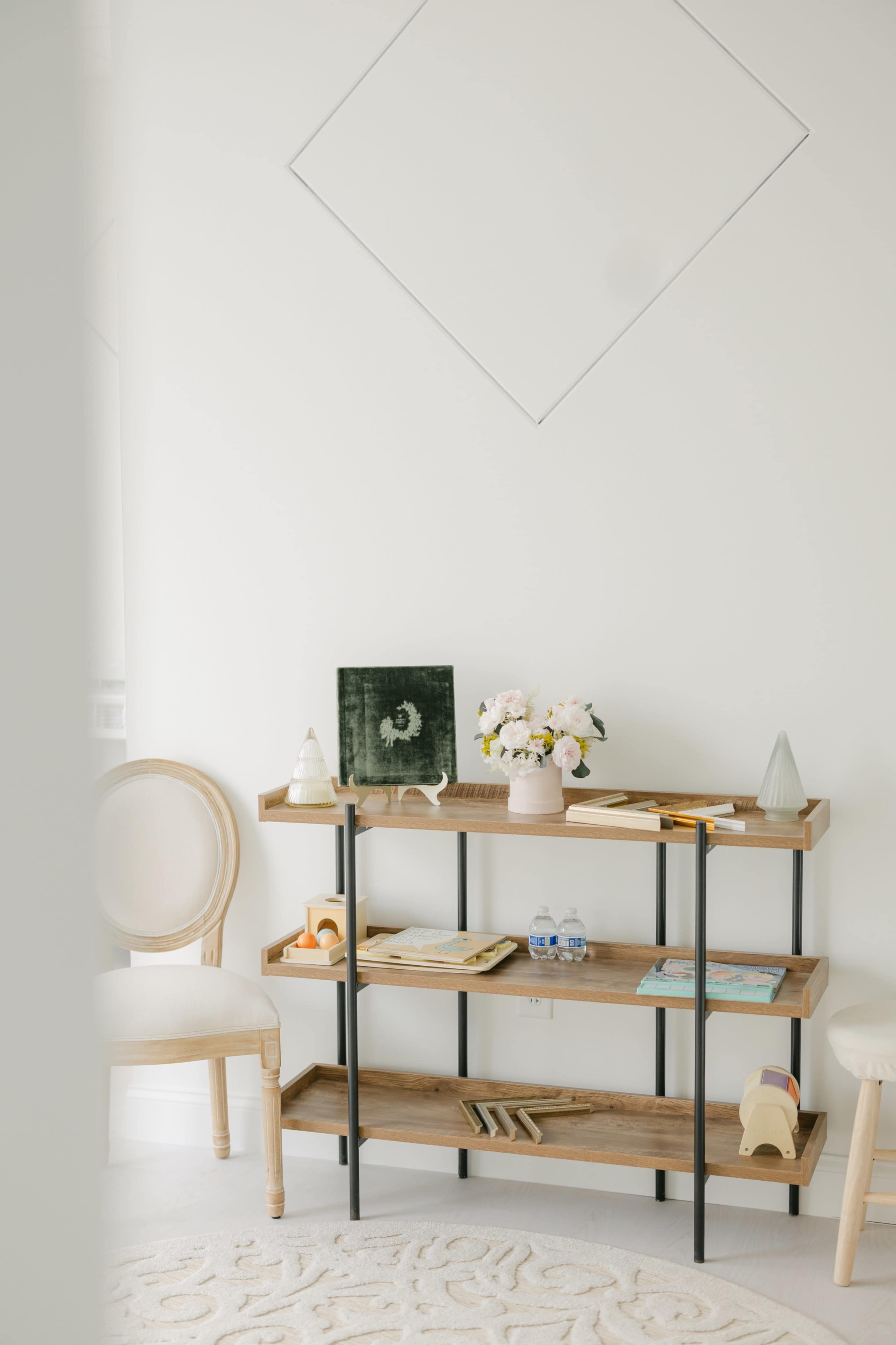 The image shows a wooden shelf made of planks and metal, holding books, decorations, and a vase of flowers, with a chair nearby and a diamond-shaped artwork on the wall.