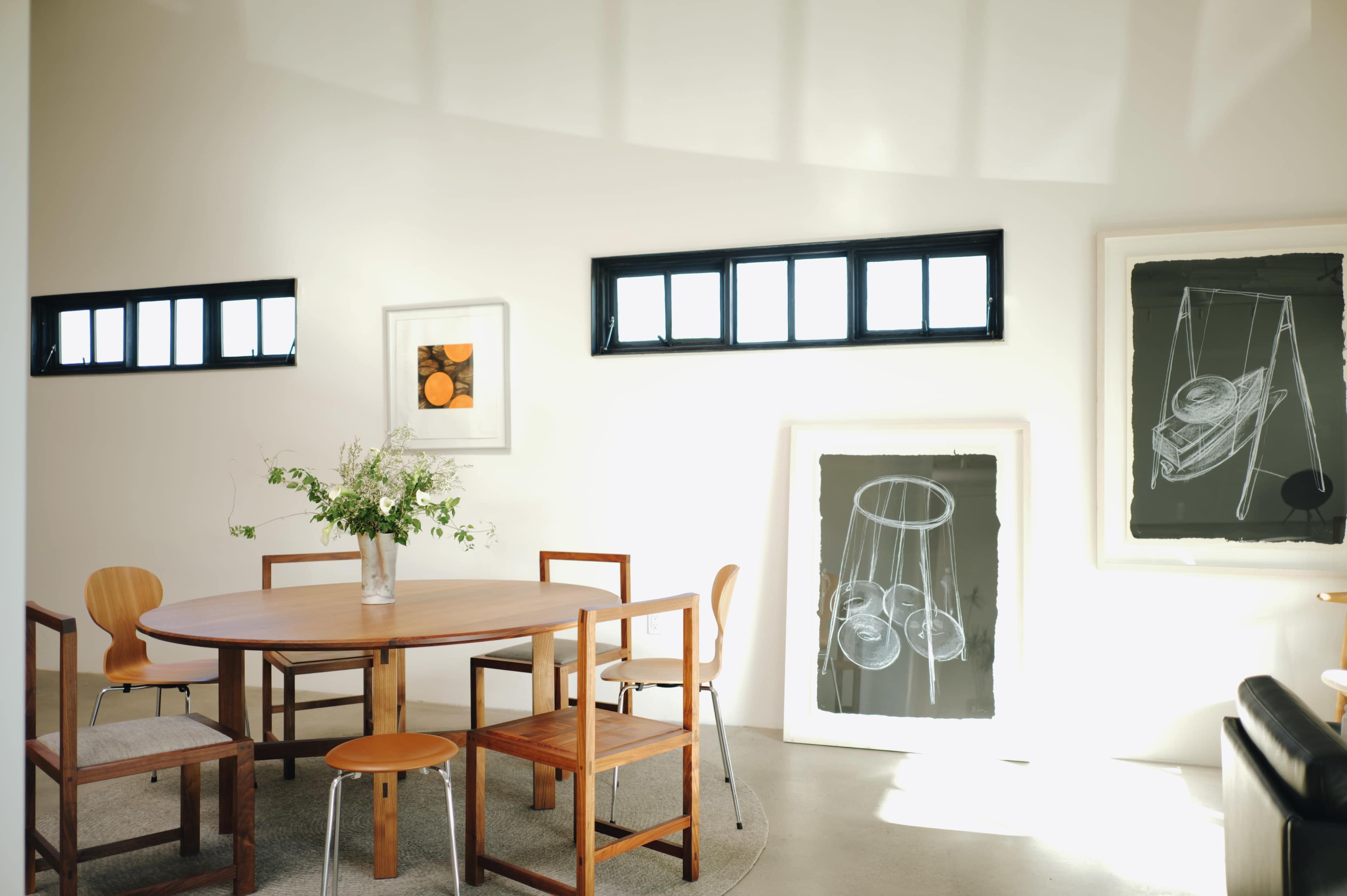A round wooden table is surrounded by wooden chairs in a bright, minimalistic room with art on the walls and large windows letting in natural light.
