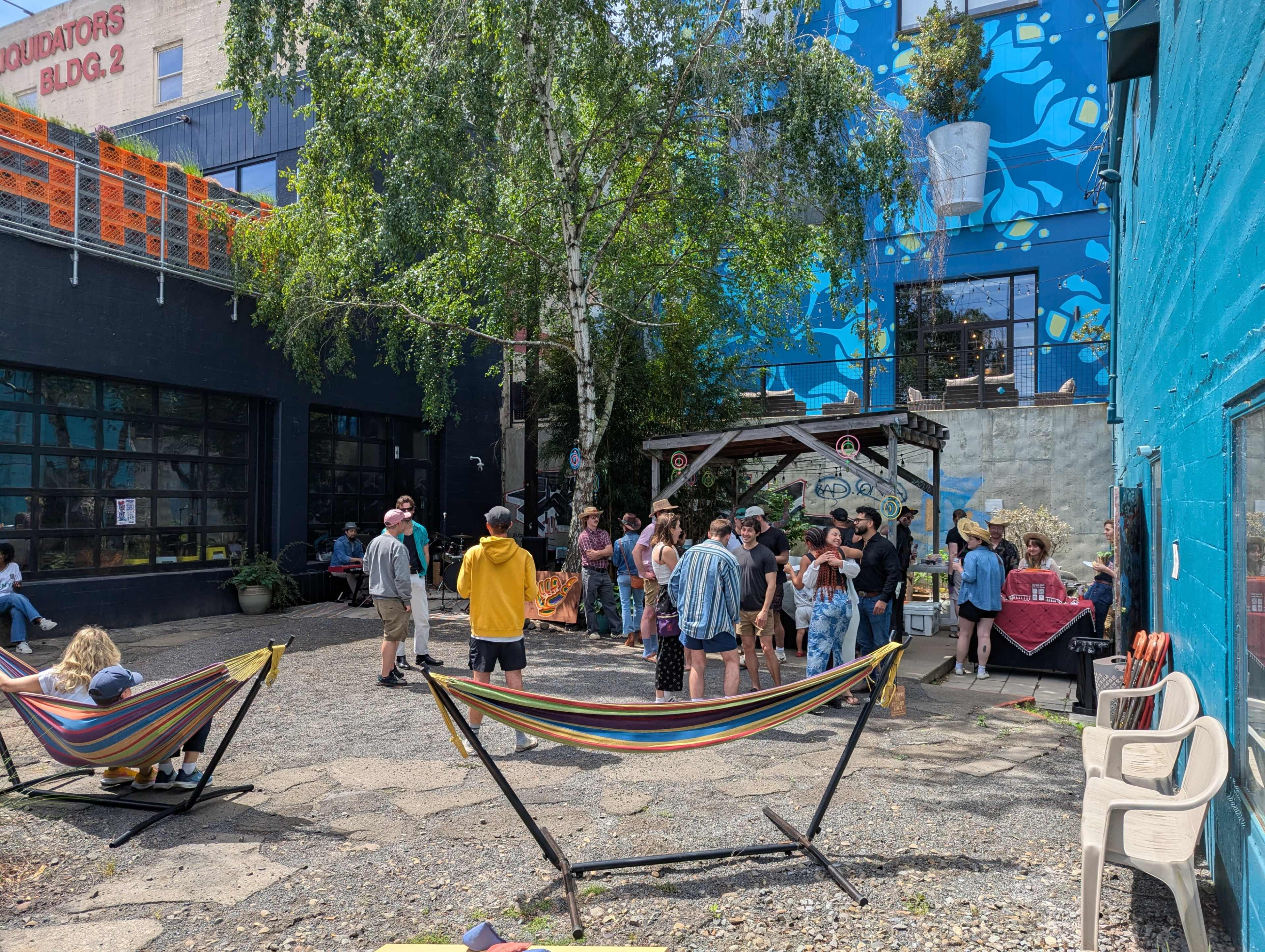 A group of people gathers in an outdoor courtyard surrounded by colorful murals, hammocks, and various seating arrangements.