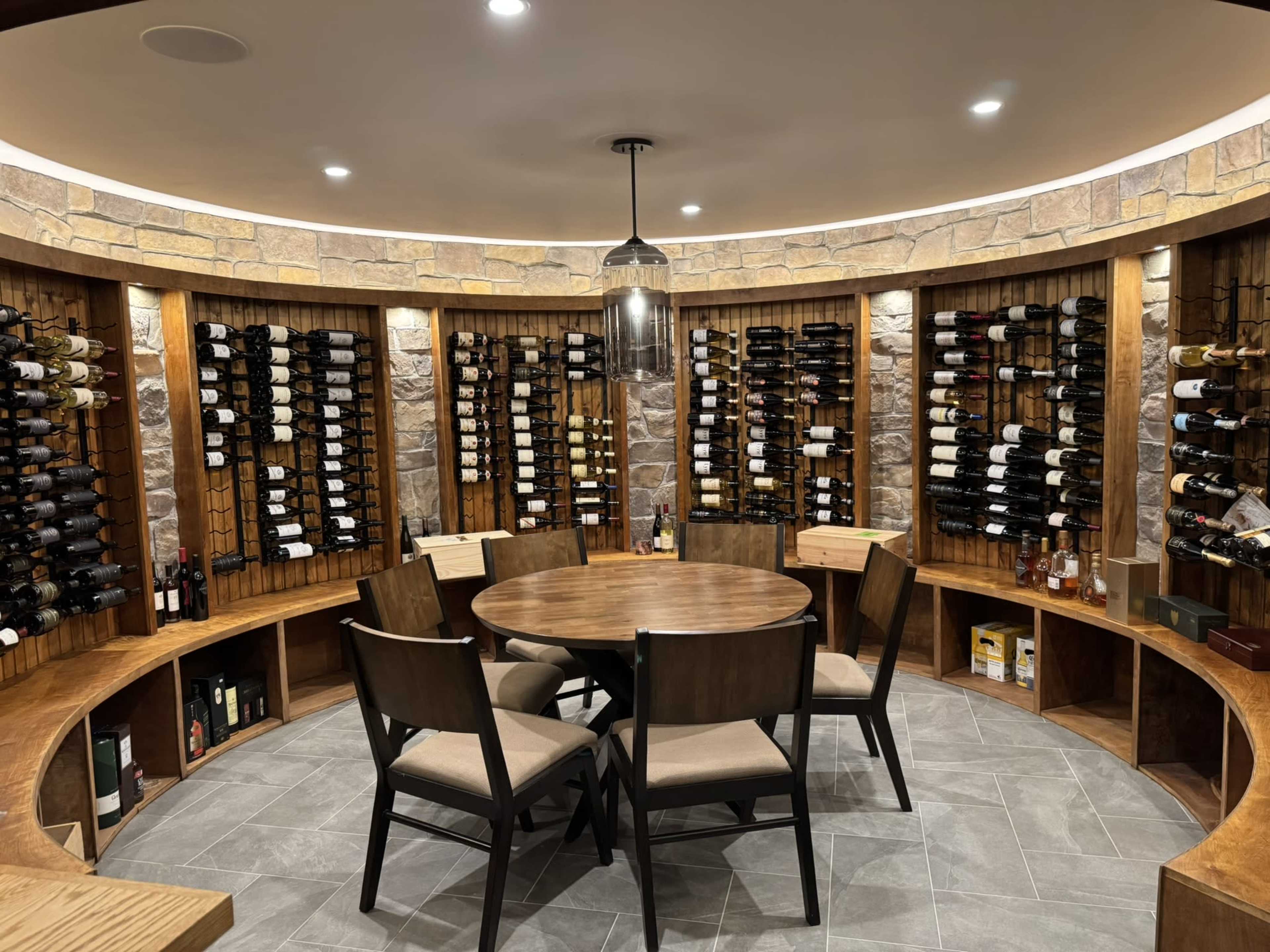 The image shows a circular wine cellar with stone walls, wooden shelving filled with bottles, and a round table surrounded by four chairs.