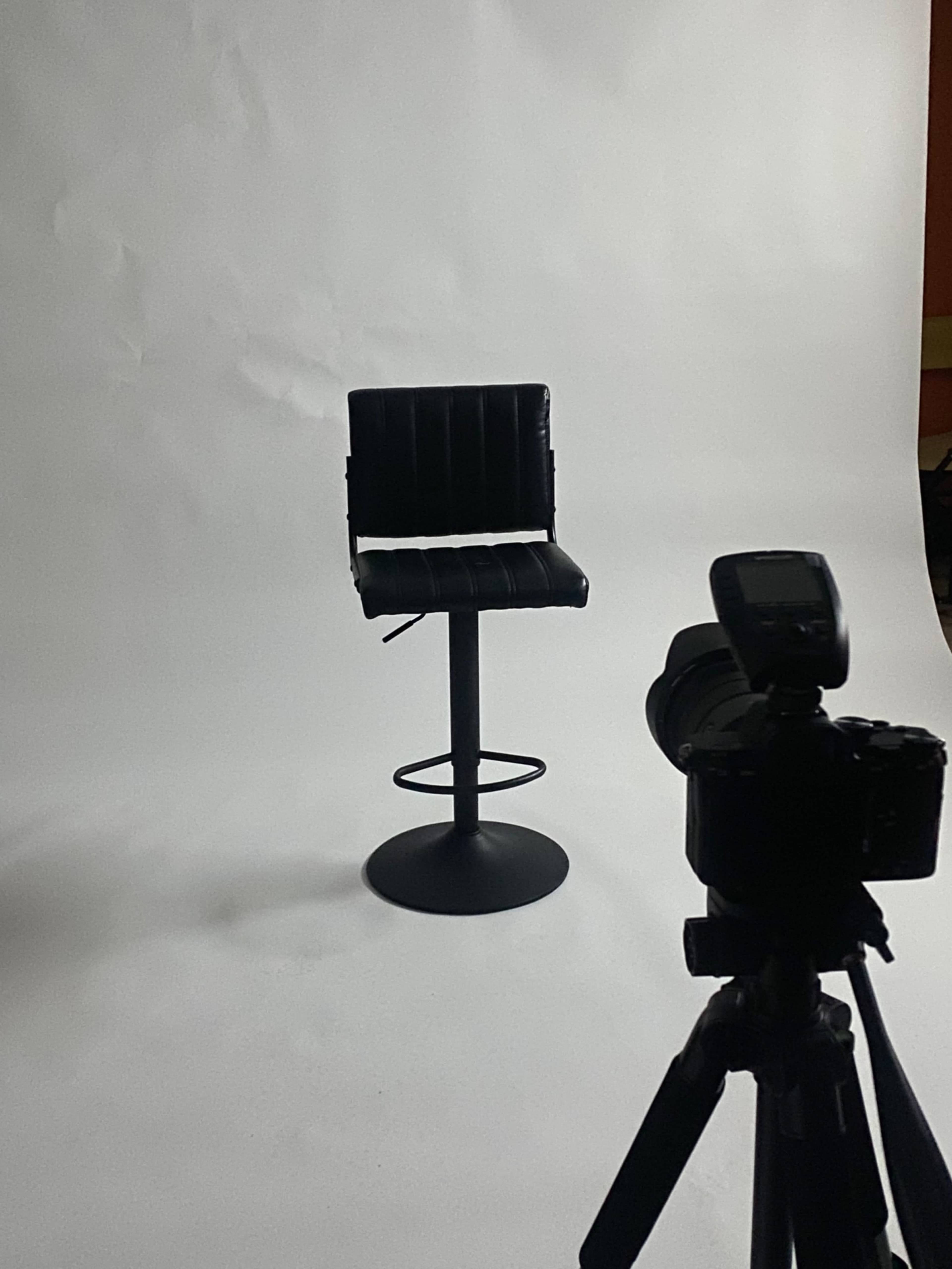 A black bar stool sits on a white backdrop in front of a camera.