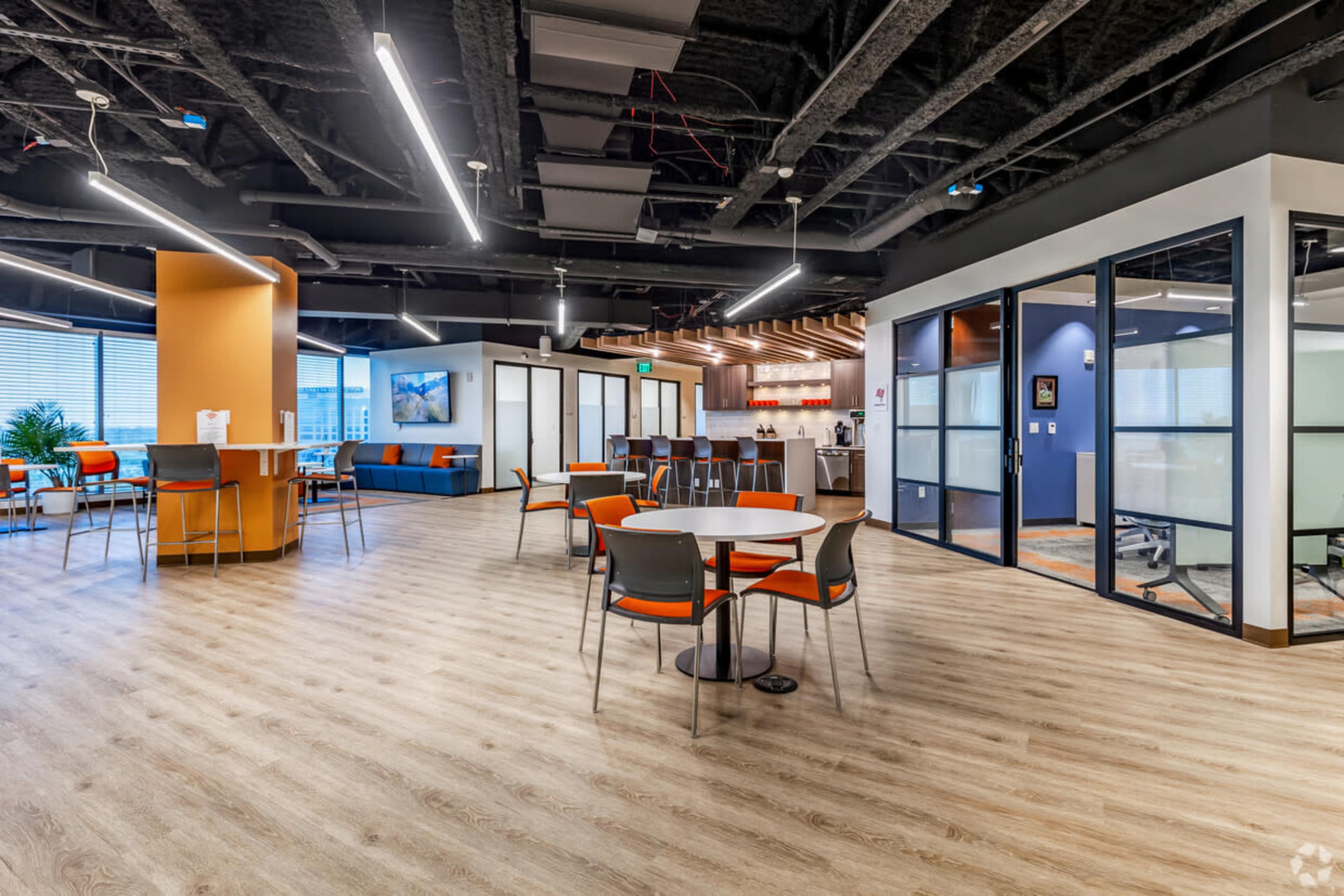 The image shows a modern office space featuring tables and chairs, an open layout with multiple seating areas, and large windows allowing natural light.