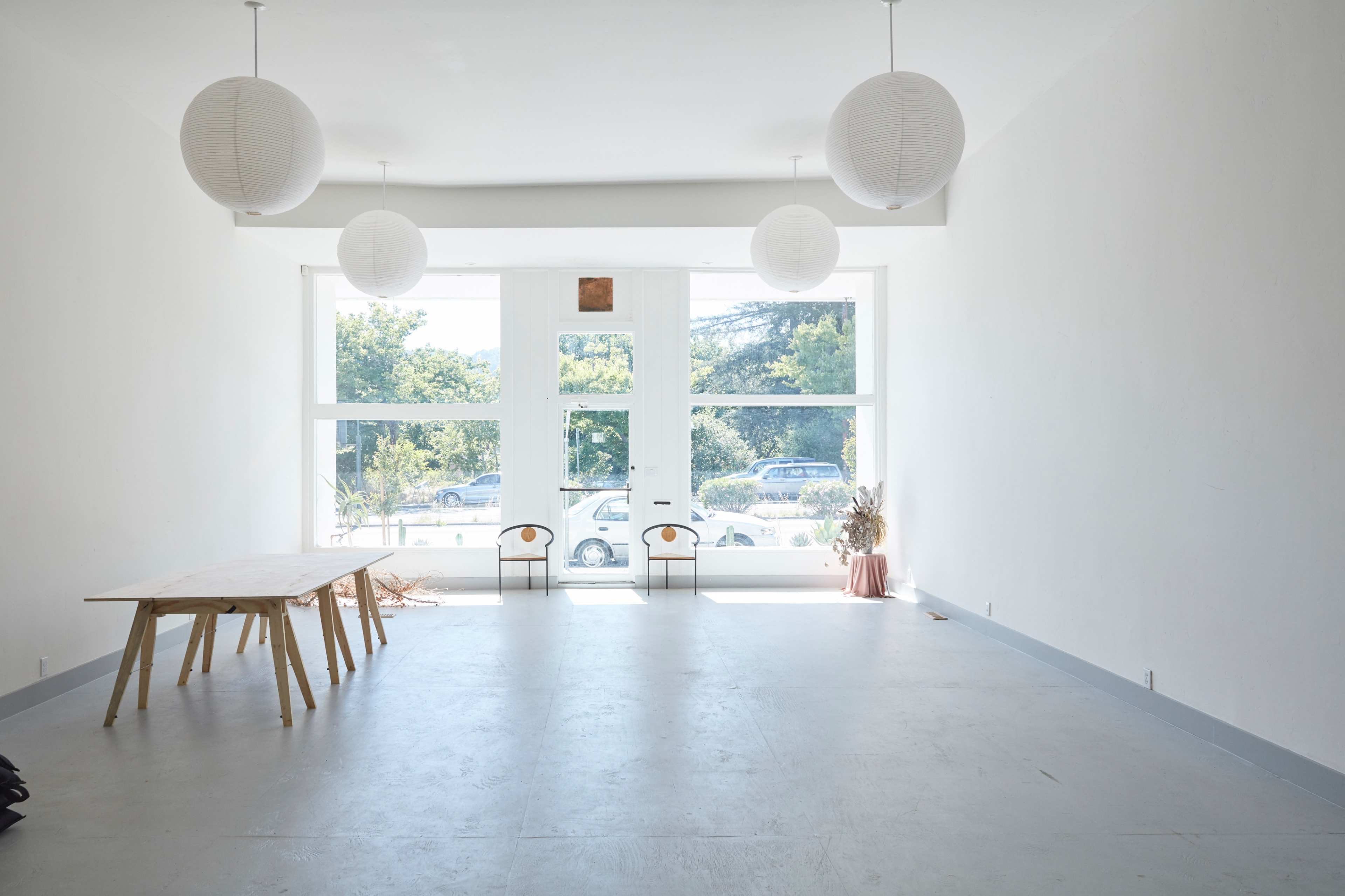 A spacious, minimalist room with large windows, a wooden table, and three paper lanterns hanging from the ceiling.