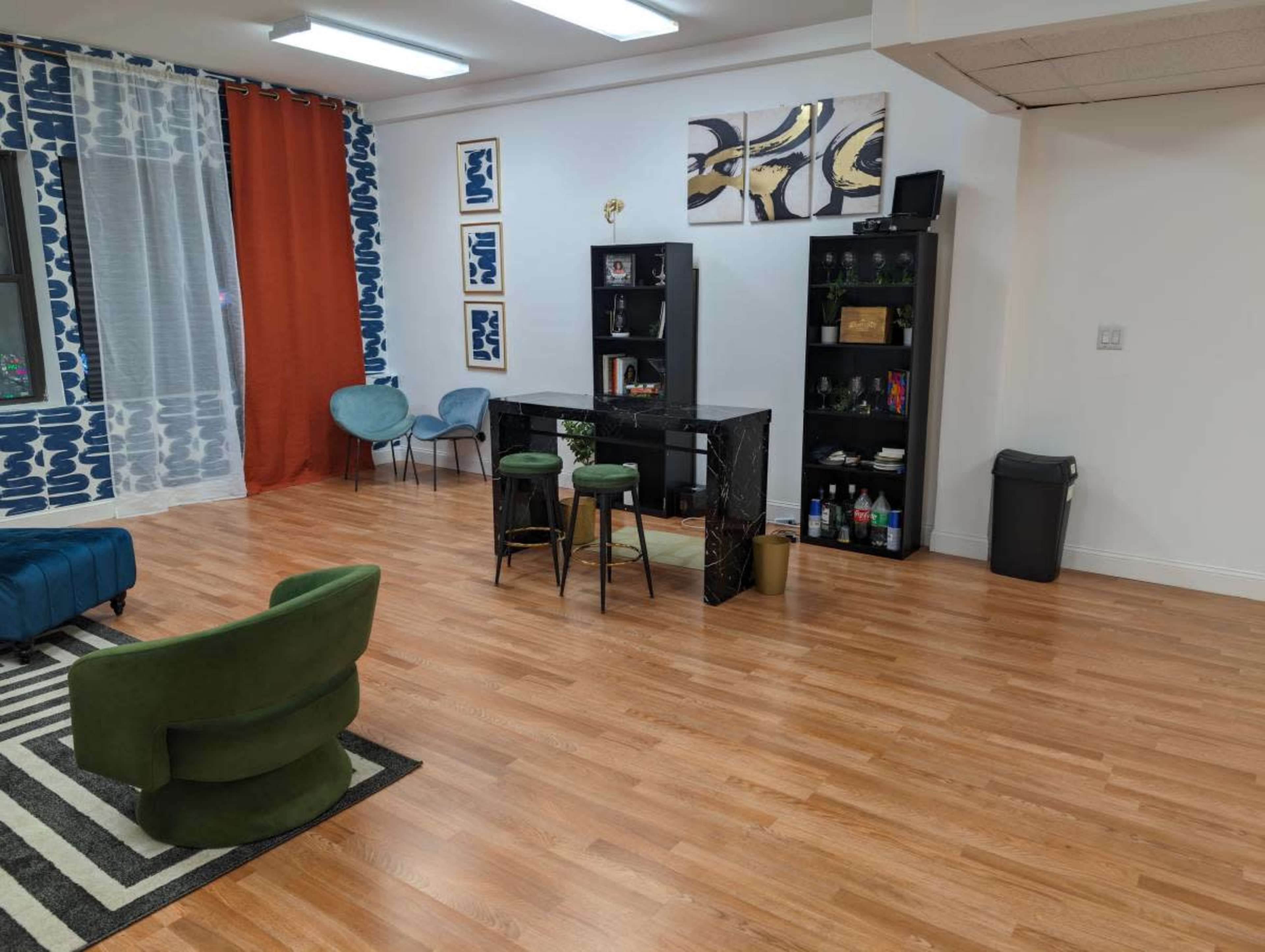 The room features a hardwood floor with a green chair, a blue sofa, a black shelf displaying decor, and a small black table with two stools, surrounded by windows and patterned curtains.
