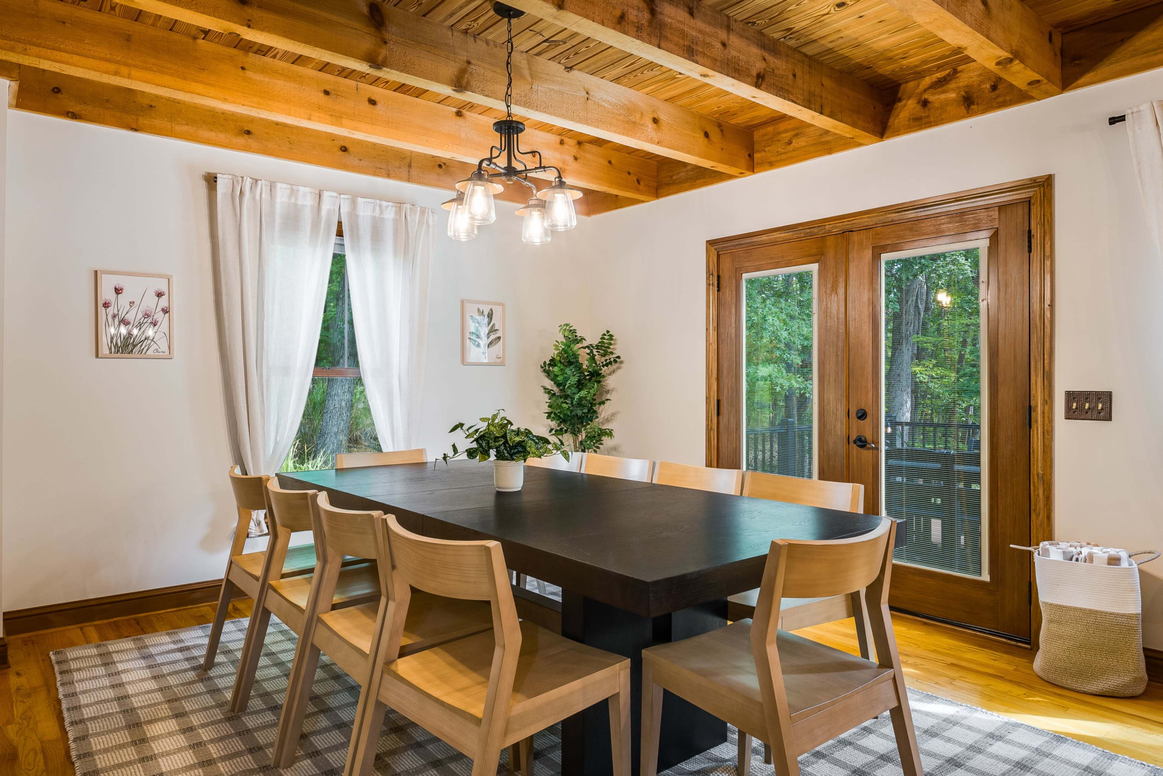 A modern dining room features a large dark table surrounded by wooden chairs, with a window and sliding glass doors letting in natural light.