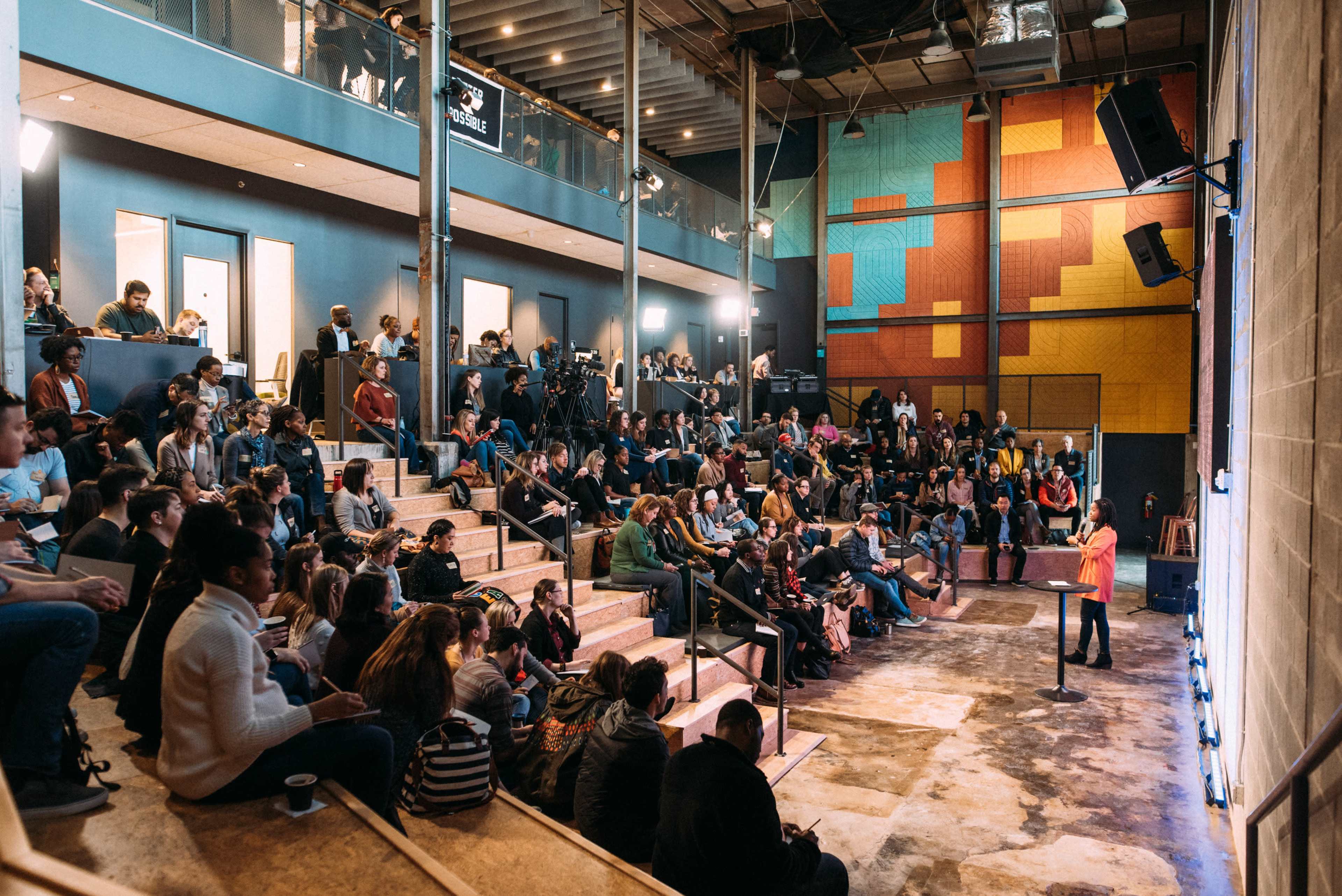 A speaker presents to a large audience seated in a tiered venue with colorful walls and stage lights.