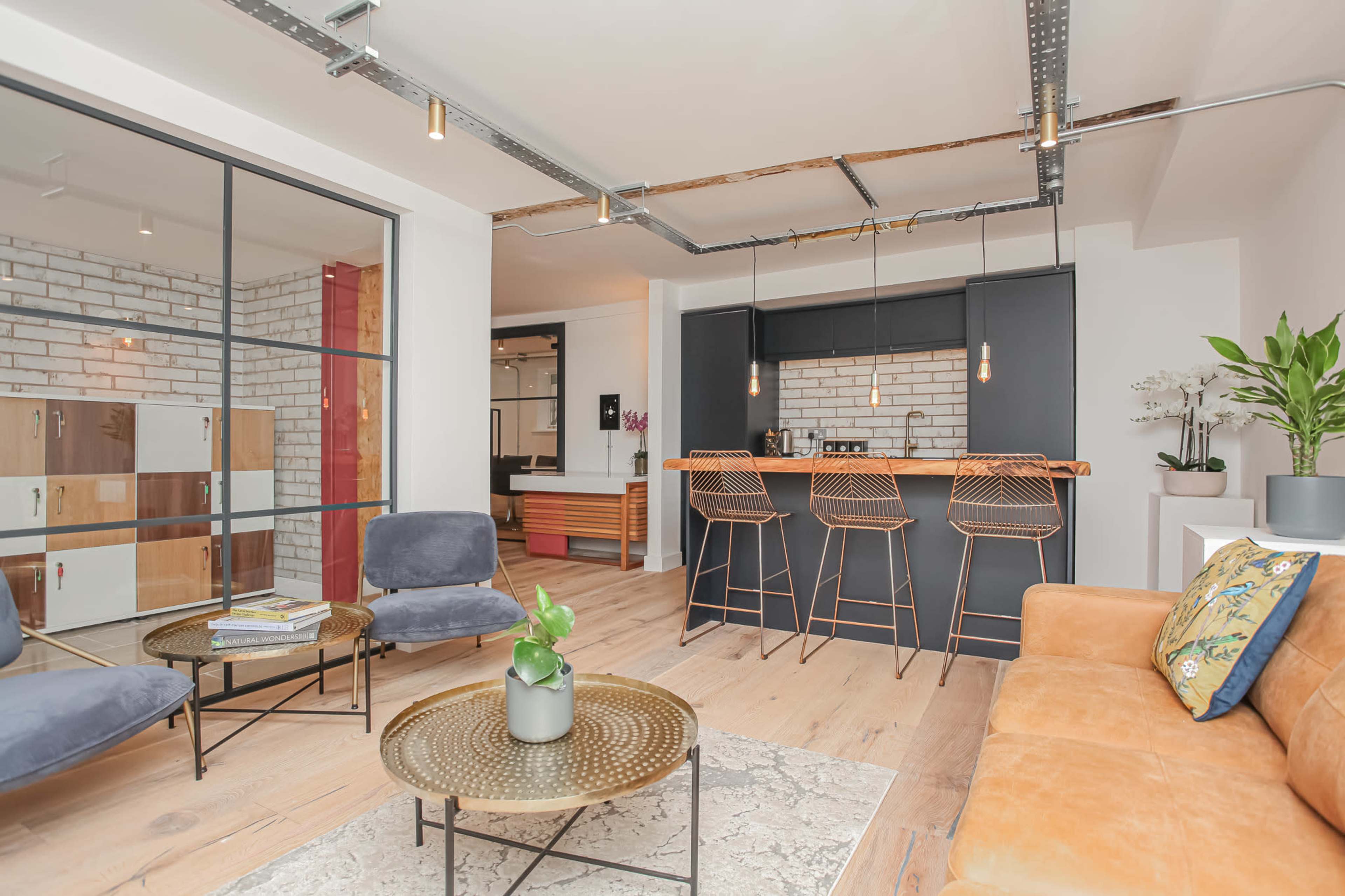 The image depicts a modern living space with a lounge area featuring a brown sofa, gray chairs, and a round coffee table, alongside a kitchen bar with high stools and a brick backsplash.