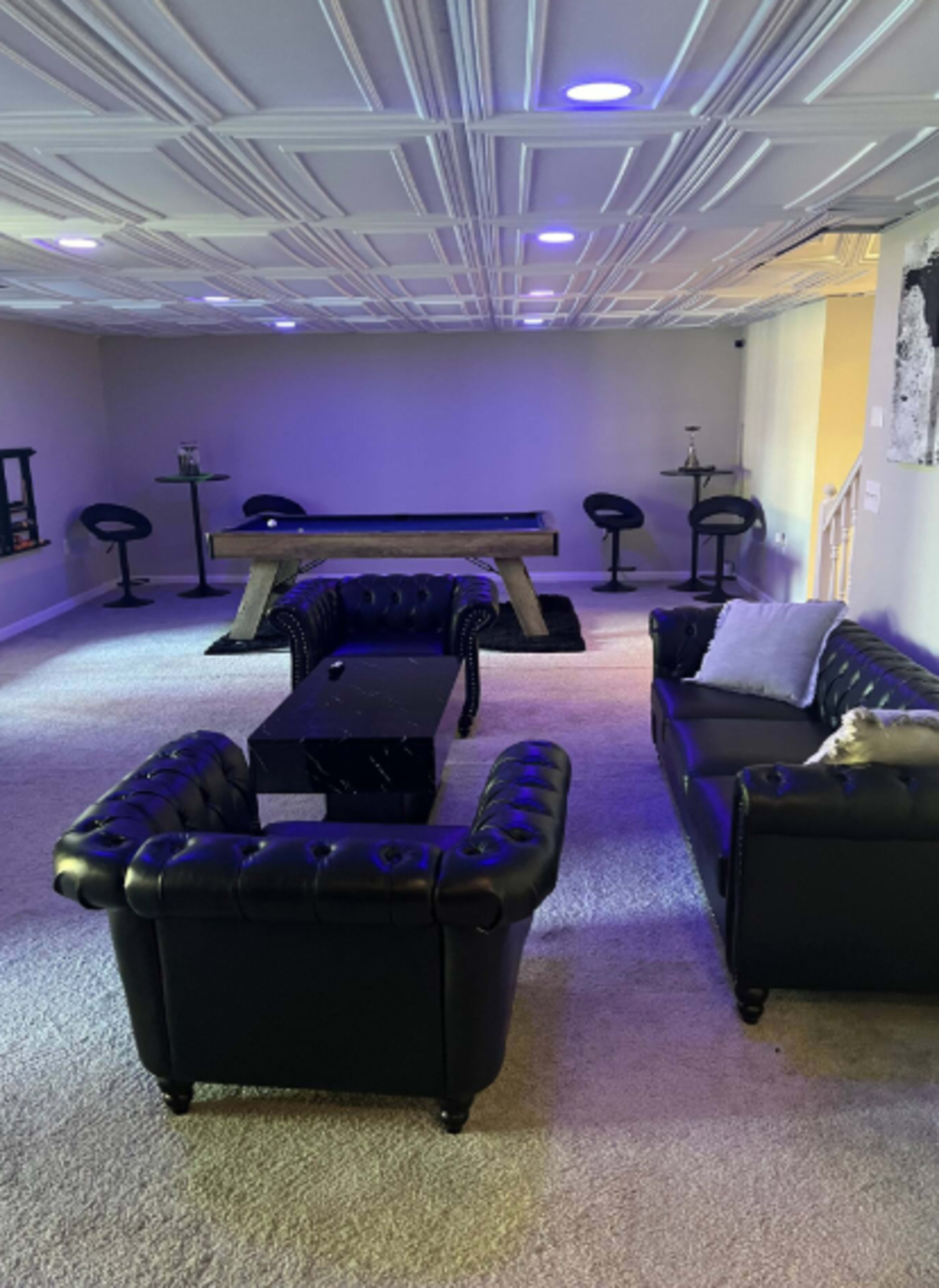 The image shows a spacious room with a pool table, black leather furniture, and bar-style seating under soft lighting.
