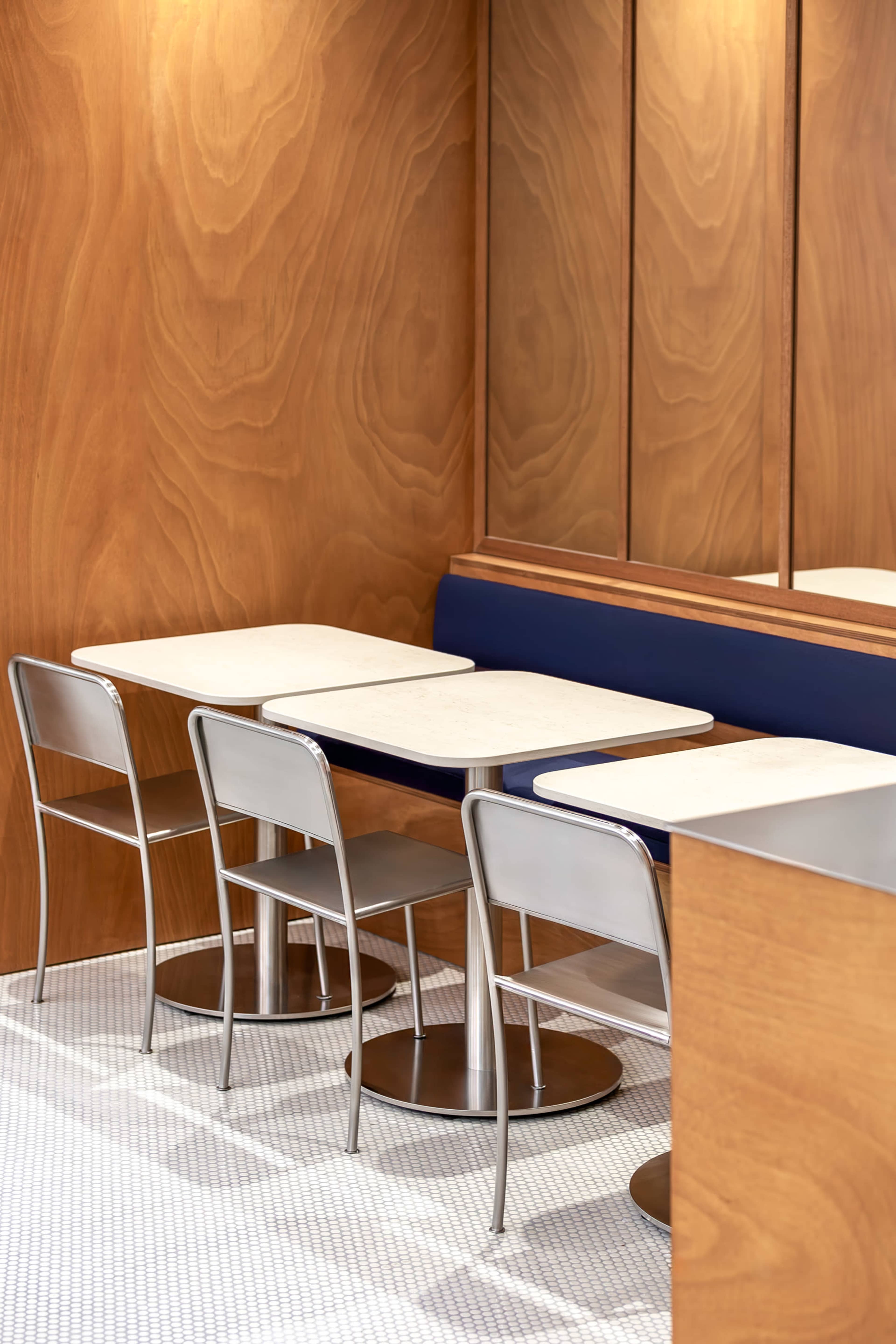 A minimalistic dining area features two marble-topped tables and metal chairs against a wood-paneled wall with a blue seating area.