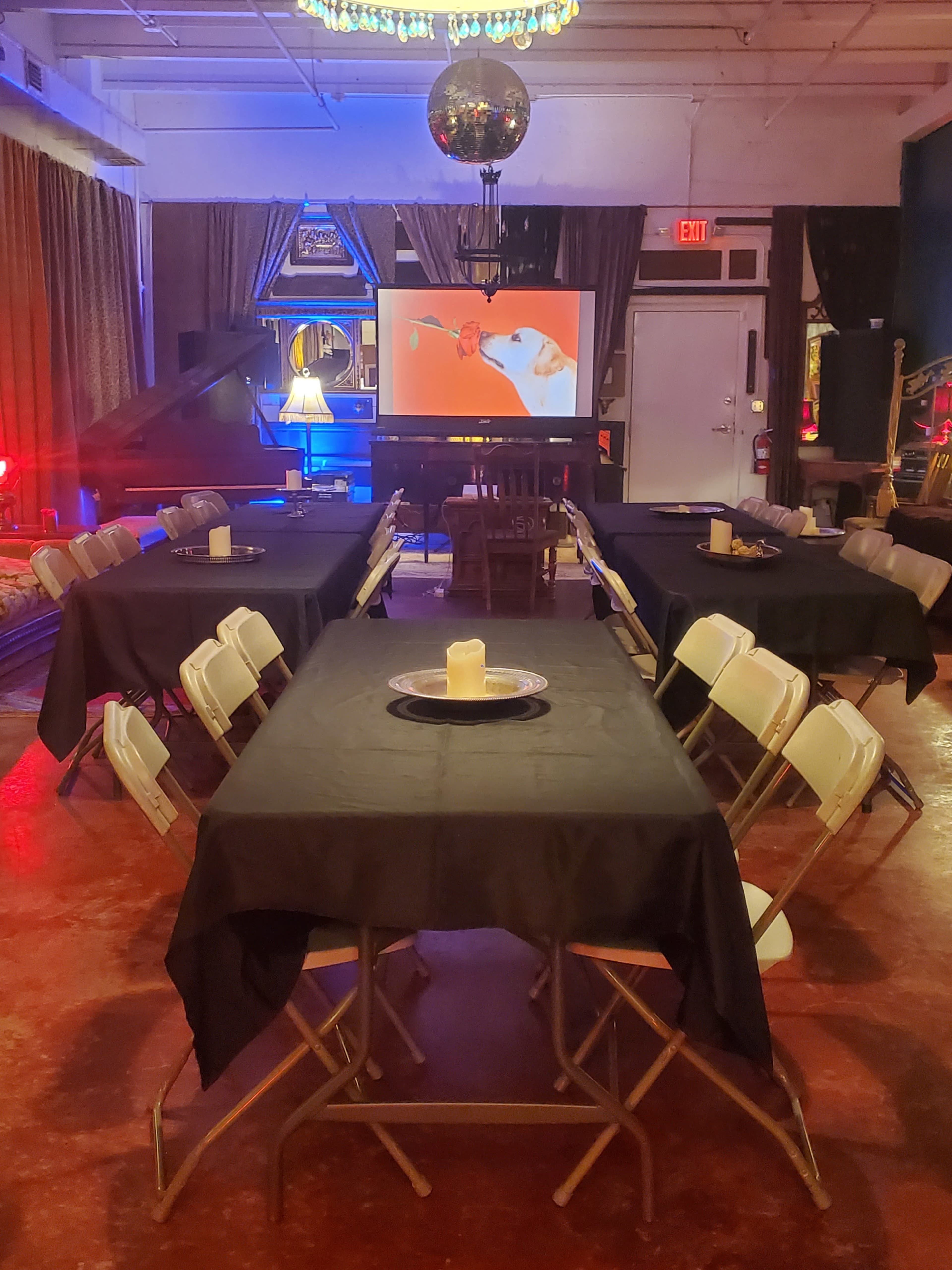 A room with several tables covered in black cloth, arranged in a U-shape, features a screen displaying a film and is illuminated with colorful lights.