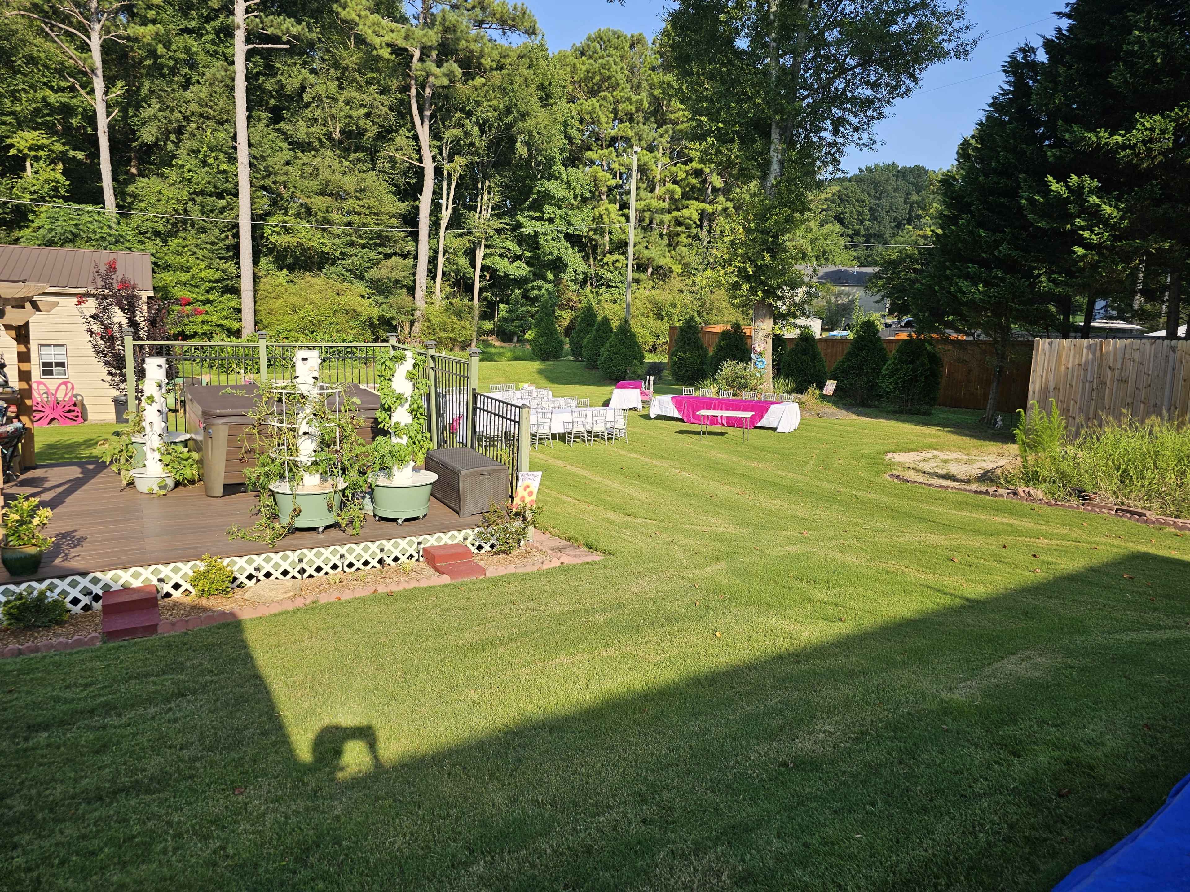 The image shows a landscaped backyard featuring a wooden deck with potted plants, a grassy area, and several tables set up for an event.