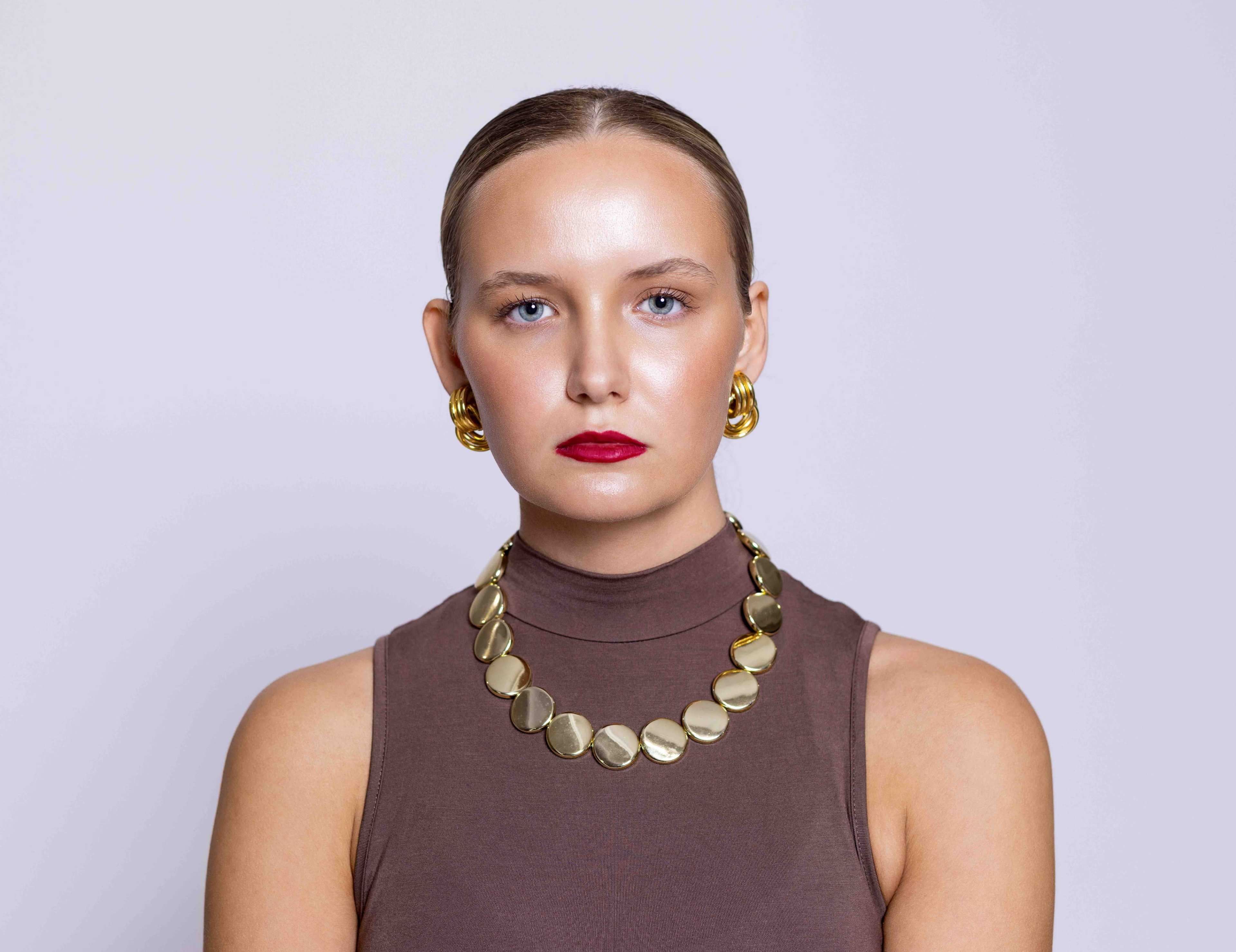 A woman with a sleek hairstyle wears a brown sleeveless top and gold jewelry, facing the camera against a light background.
