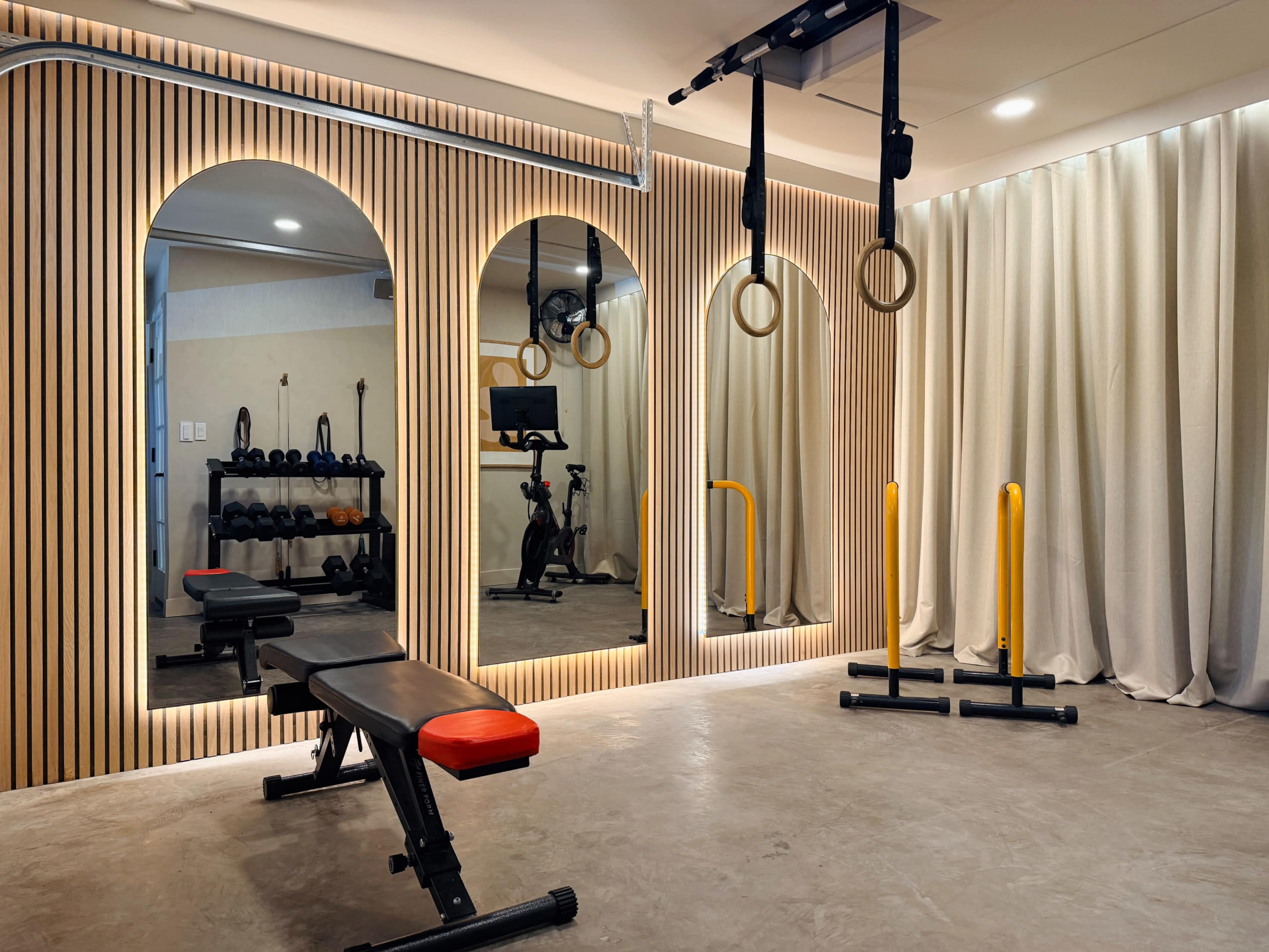 The image shows a neatly arranged home gym featuring a weight bench, exercise rings, and a mirror with ambient lighting, along with a collection of dumbbells and a stationary bike in the background.