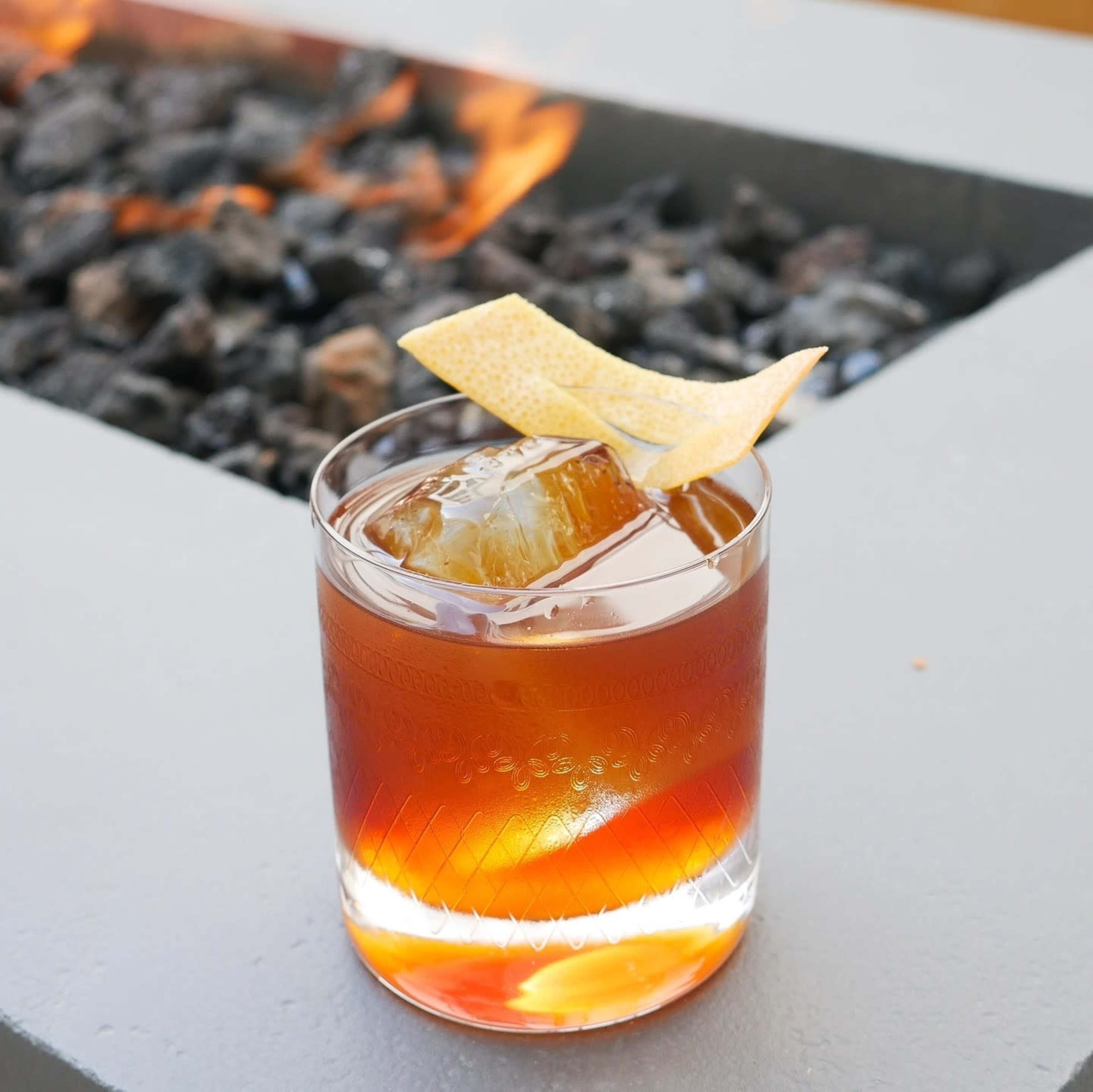 A clear glass filled with a brown cocktail and a large ice cube sits on a light gray surface next to a fire pit with flickering flames.