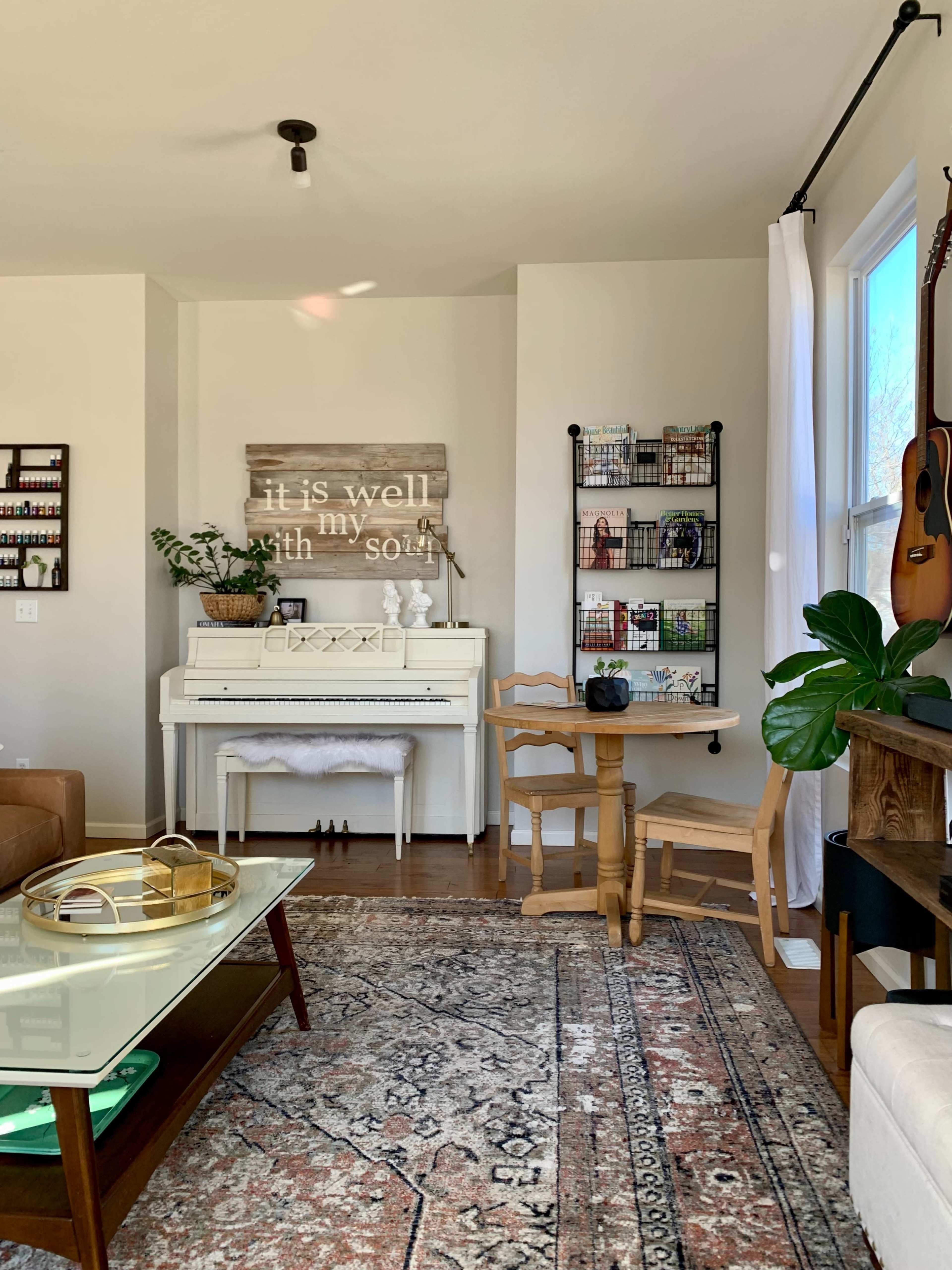 A cozy interior featuring a white piano, a small dining table, a bookshelf, and a decorative wall sign that reads "it is well with my soul."