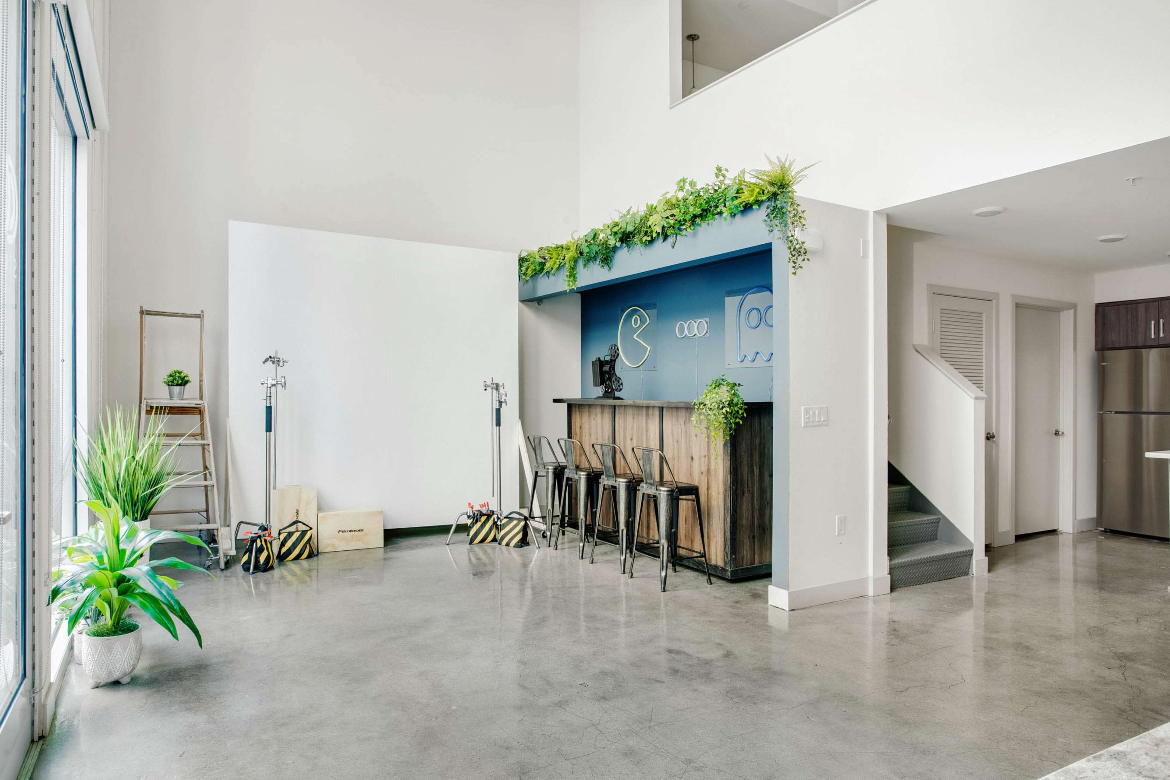 A modern and spacious interior with a wooden bar, metal stools, and potted plants, alongside a staircase and kitchen appliances in a well-lit room.