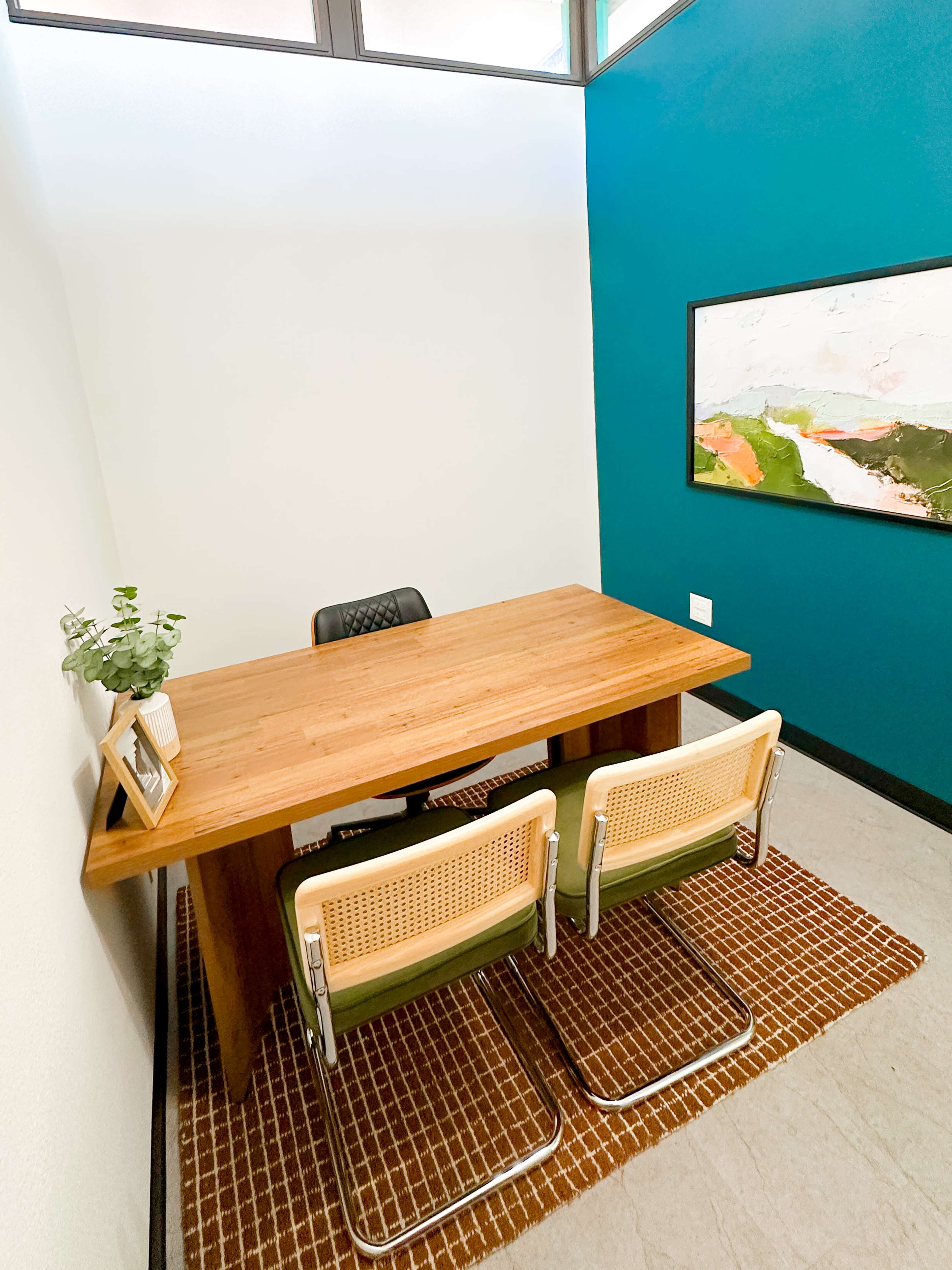 A small office space features a wooden desk, two chairs with wicker seats, and a colorful abstract painting on the wall.