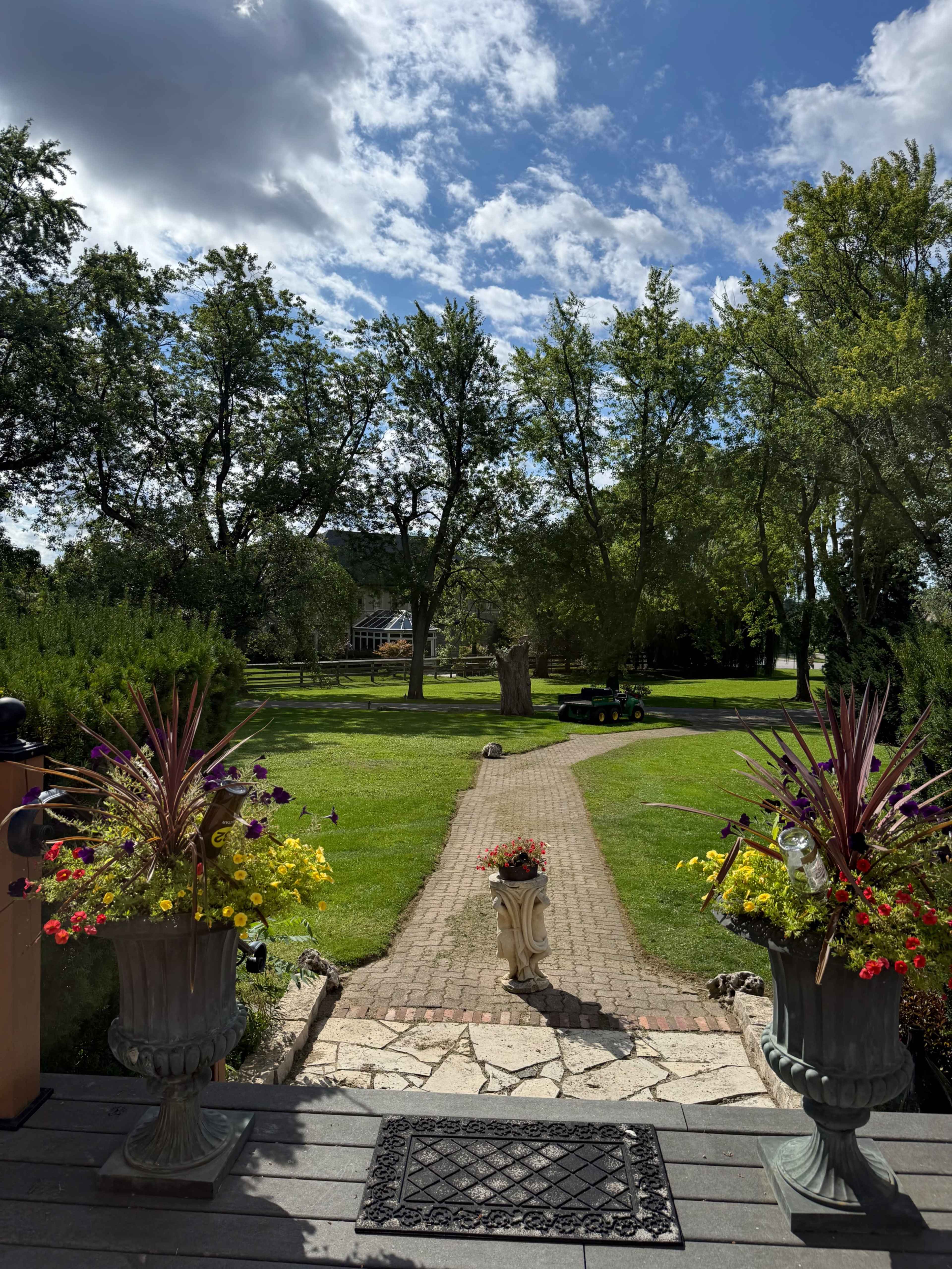 A pathway lined with flower pots leads through a green lawn surrounded by trees under a partly cloudy sky.