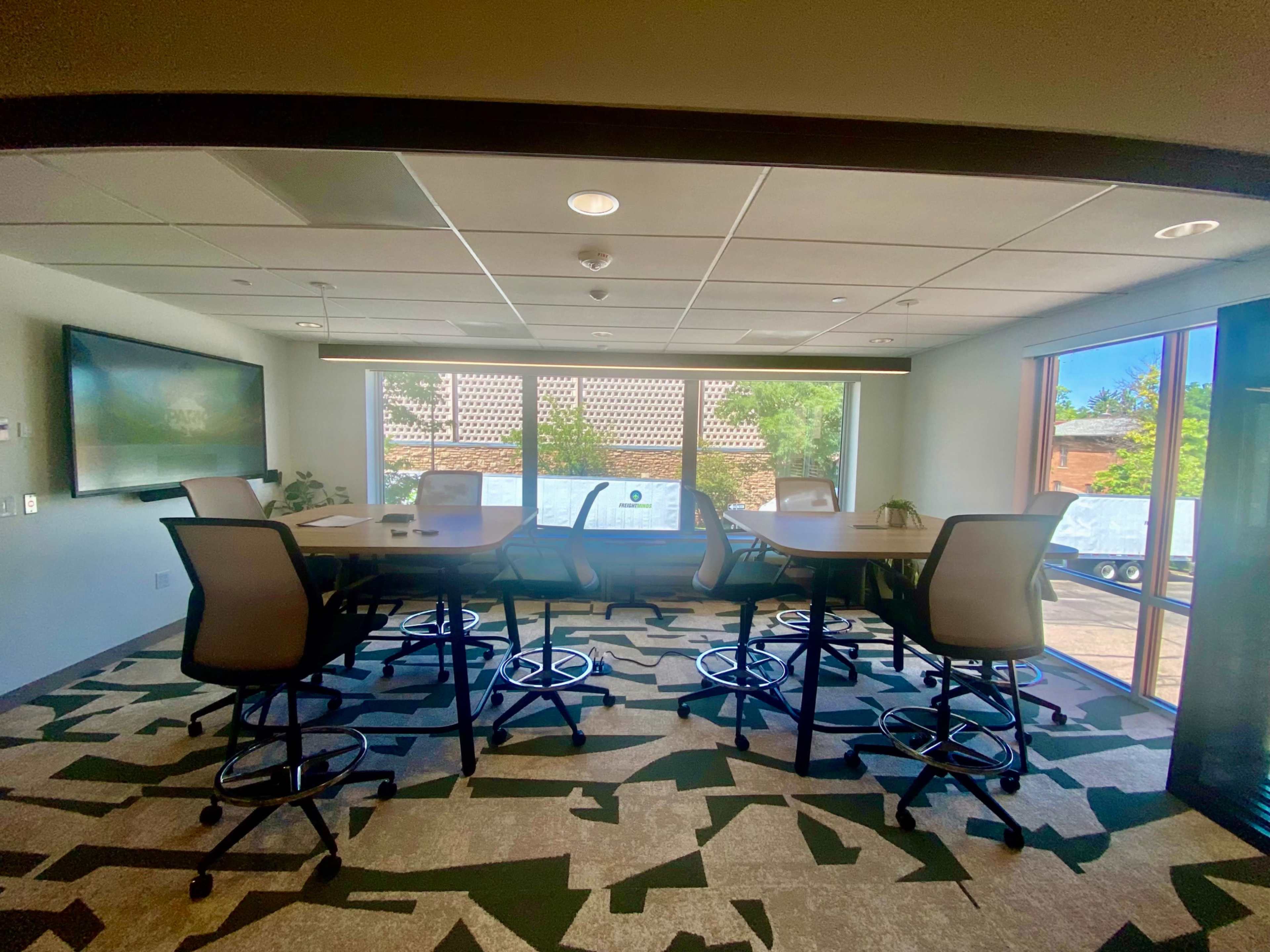 A modern conference room features a large table surrounded by high-backed chairs, with windows that provide natural light and views of the outdoors.