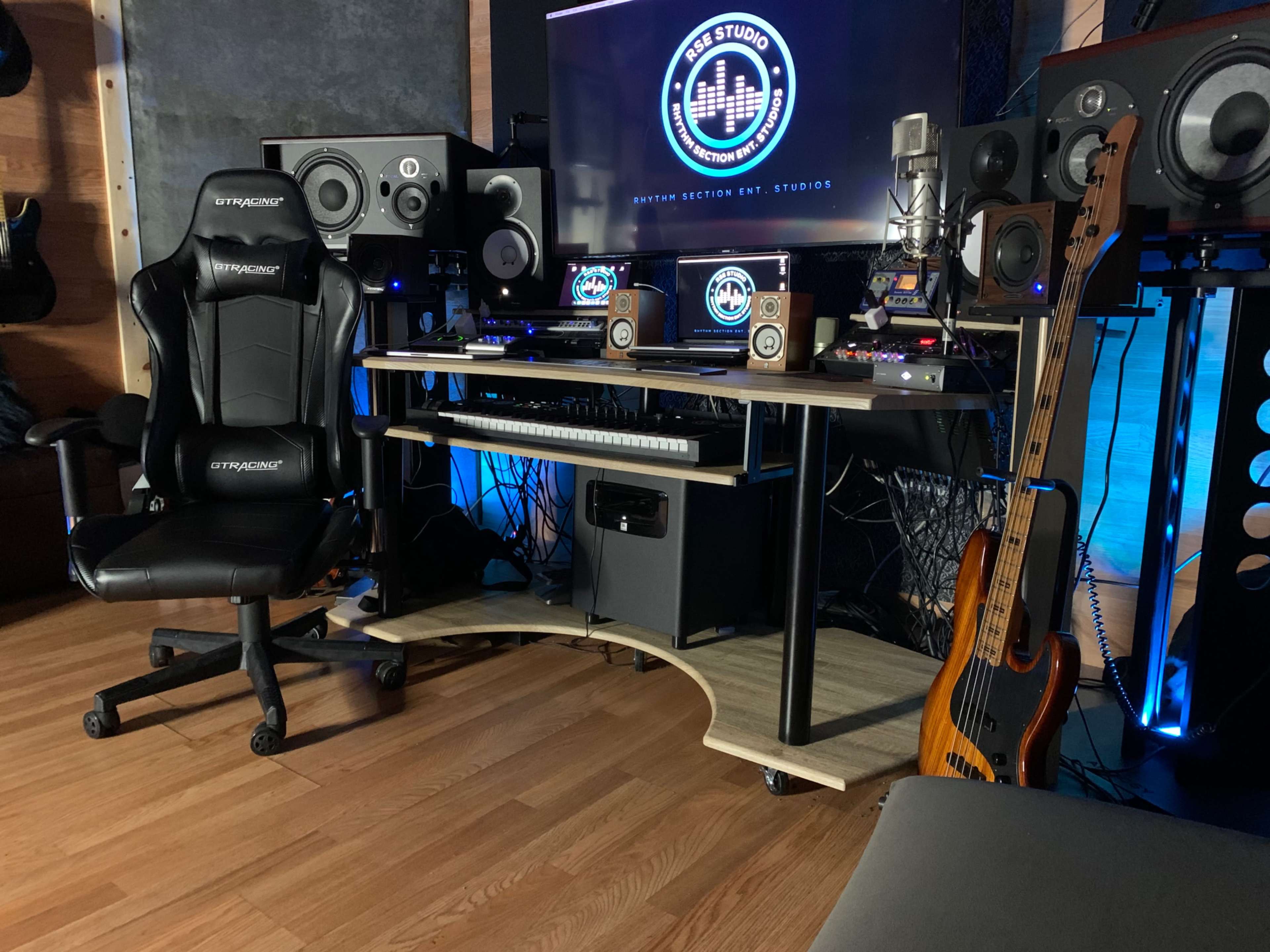 A recording studio setup features a desk with audio equipment, monitors, a guitar, and a gaming chair.
