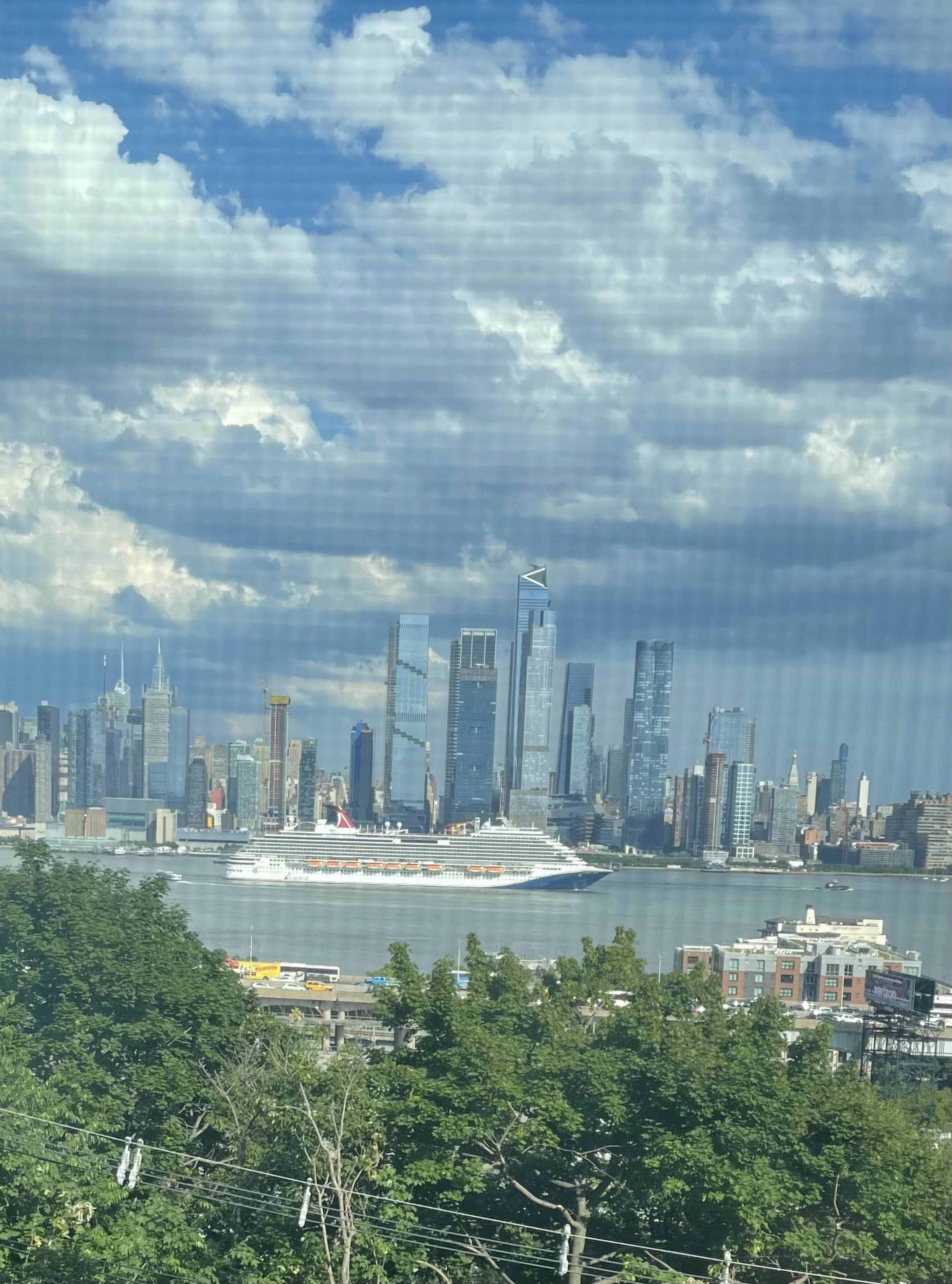 A large cruise ship is docked in the water with a skyline of tall buildings and clouds in the background.