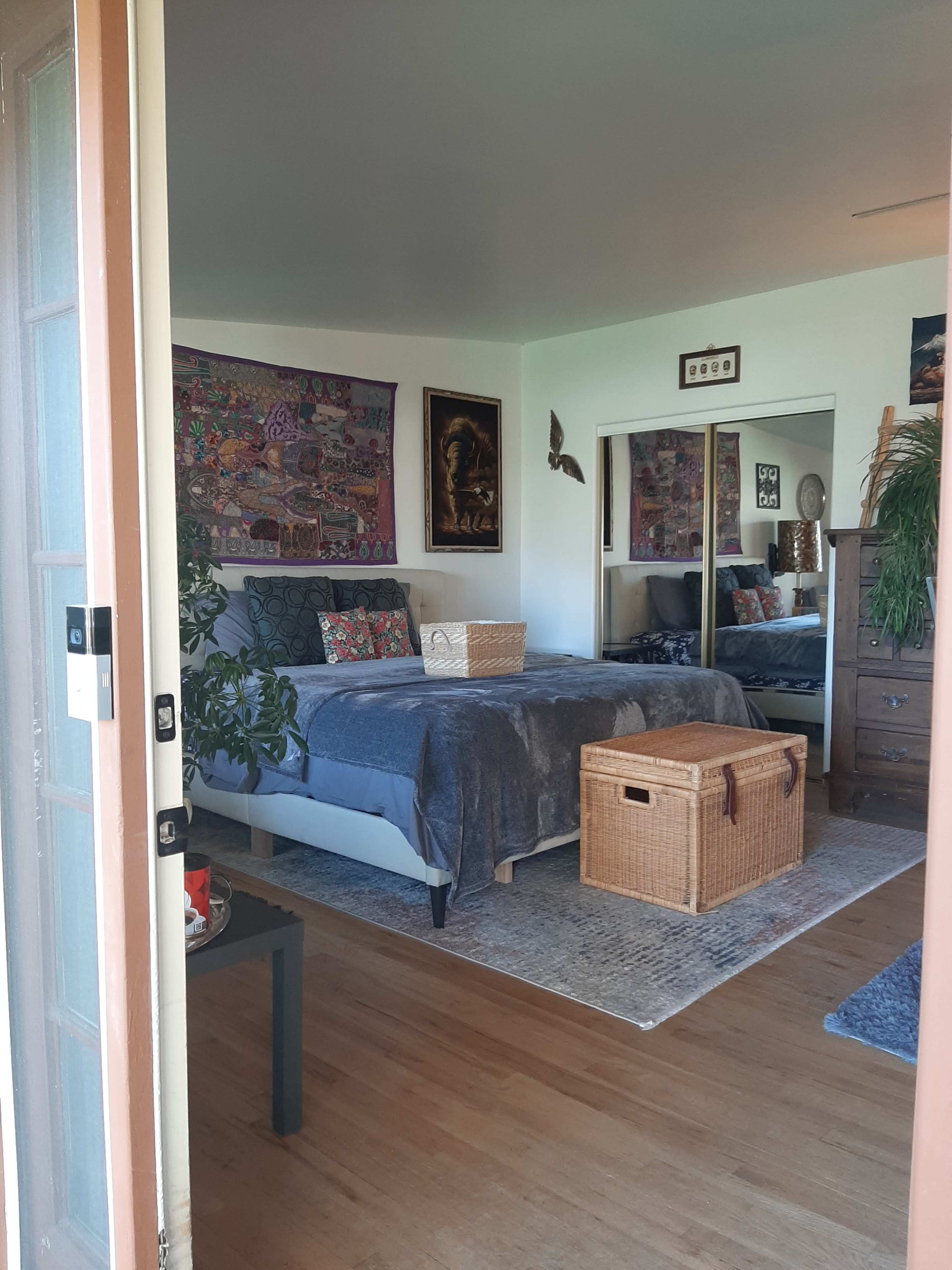 A cozy bedroom with a bed, decorative wall art, a large mirror, and a woven storage basket at the foot of the bed.