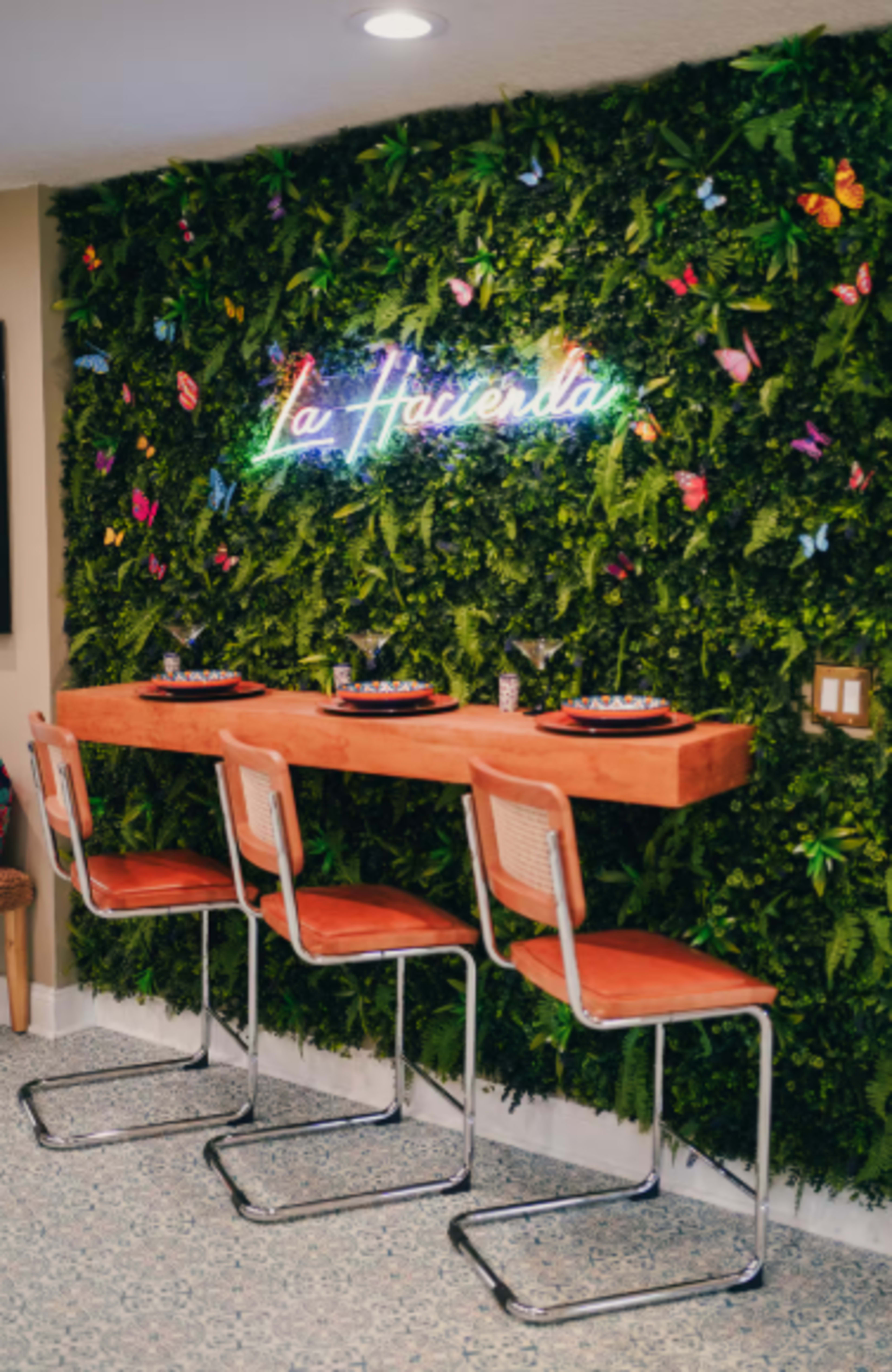 The image features a bar area with a wooden counter and three chrome stools in front of a vibrant green wall adorned with artificial foliage and colorful butterflies, illuminated by a neon sign that reads "La Hacienda."